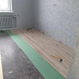 Daniel Borzęcki - Układanie paneli podłogowych w pokoju: widoczne zielone podkłady izolacyjne, częściowo ułożone jasne panele, kątownik stolarski i ołówek na panelach, surowa podłoga i ściany w trakcie remontu.