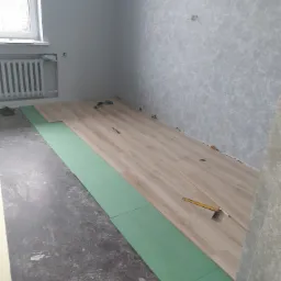 Układanie paneli podłogowych w pokoju: widoczne zielone podkłady izolacyjne, częściowo ułożone jasne panele, kątownik stolarski i ołówek na panelach, surowa podłoga i ściany w trakcie remontu.