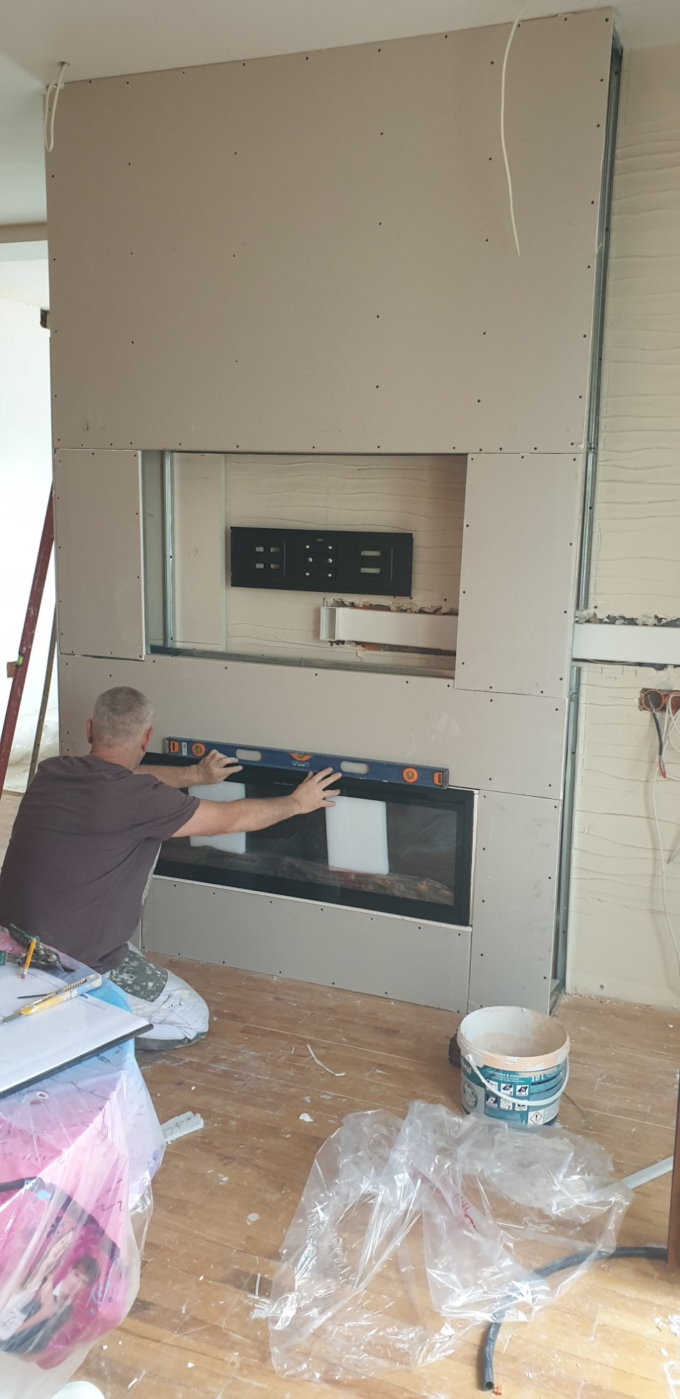 Pracownik poziomuje obudowę kominka wykonaną z płyt gipsowo-kartonowych, widoczny uchwyt do telewizora w przygotowanym otworze, surowe drewniane podłoże.