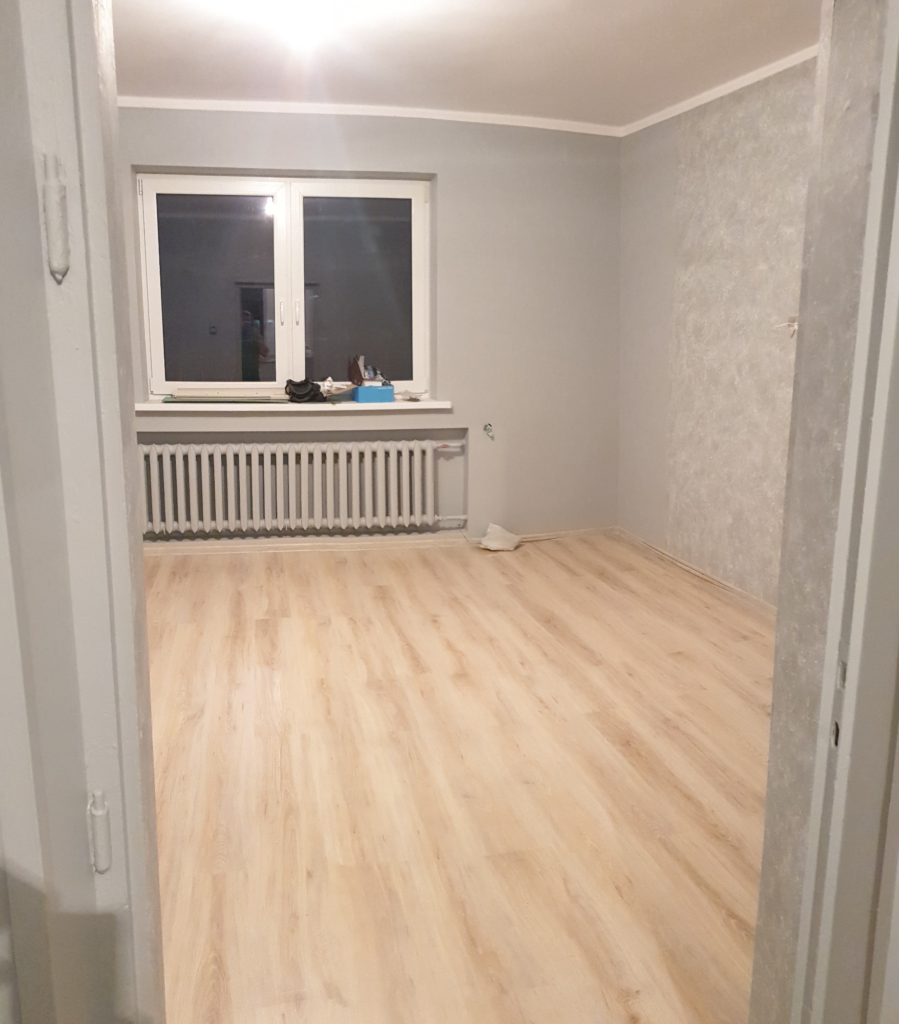 Pusty pokój po remoncie z jasnymi panelami podłogowymi, szarymi ścianami i białym grzejnikiem pod oknem, widok zza framugi drzwi.