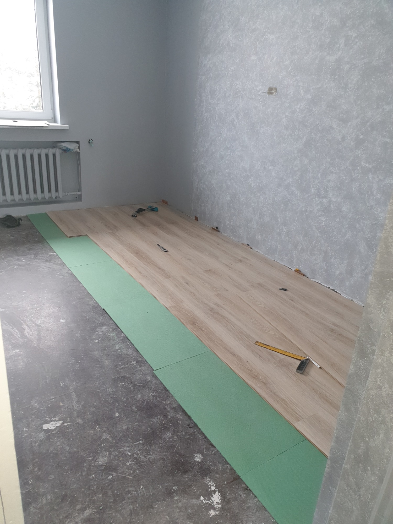 Układanie paneli podłogowych w pokoju: widoczne zielone podkłady izolacyjne, częściowo ułożone jasne panele, kątownik stolarski i ołówek na panelach, surowa podłoga i ściany w trakcie remontu.