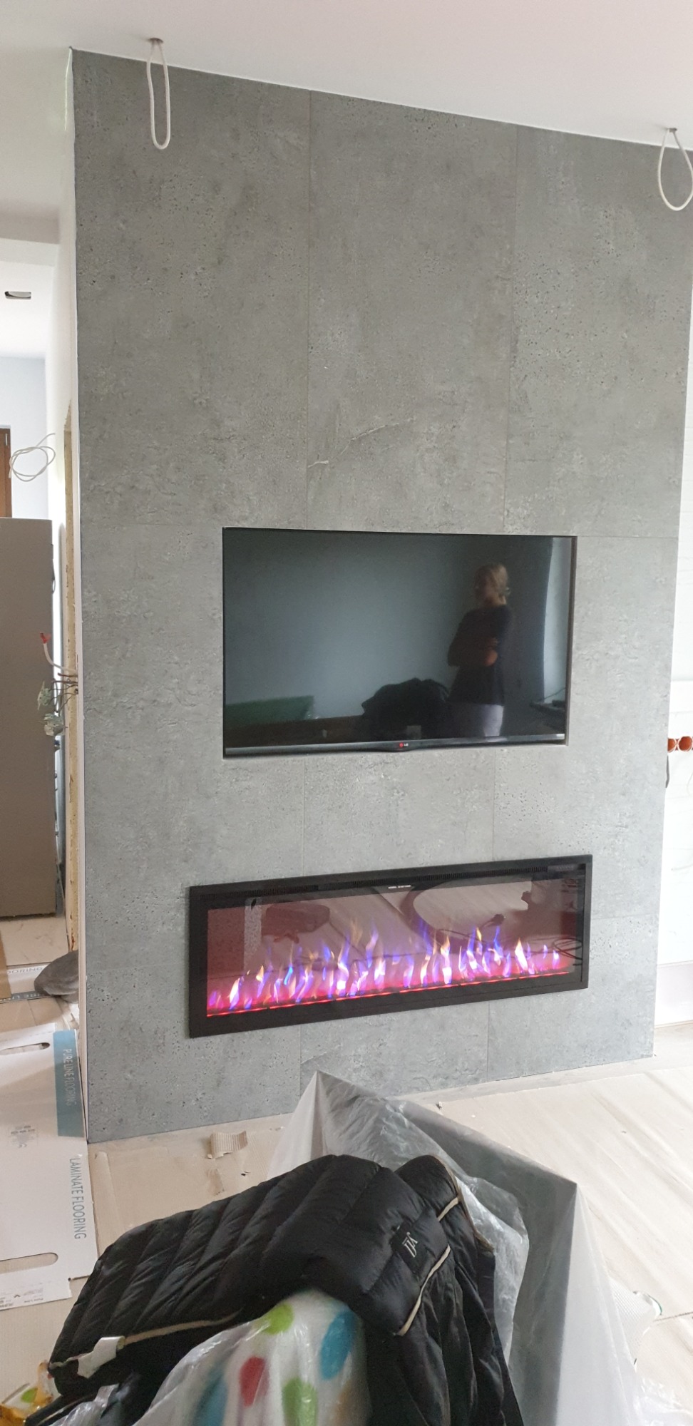 Ściana wykończona szarymi płytami imitującymi beton, z wnęką na telewizor i elektryczny kominek z efektem płomieni, widoczne kable wystające z sufitu i podłoga zabezpieczona folią malarską.