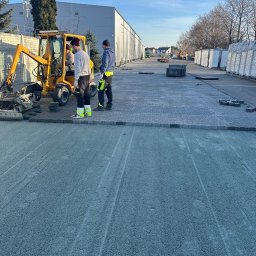 Sion-Bruk Śp. Z. O. O. - Żółta maszyna do układania kostki brukowej w trakcie pracy. Dwóch pracowników nadzoruje proces. Częściowo ułożona kostka i przygotowane podłoże. Słoneczny dzień.