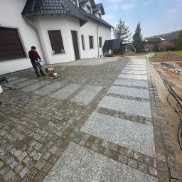 SION-BRUK SPÓŁKA Z OGRANICZONĄ ODPOWIEDZIALNOŚCIĄ - Prace brukarskie: robotnik zagęszcza wibroprasą ułożoną ścieżkę z kostki granitowej w geometryczny wzór, na tle białego domu z ciemnym dachem.