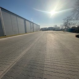 SION-BRUK SPÓŁKA Z OGRANICZONĄ ODPOWIEDZIALNOŚCIĄ - Rozległy plac wyłożony szarą kostką brukową typu 'psią kość' w słoneczny dzień. W tle widoczny budynek przemysłowy z szarej blachy trapezowej i błękitne niebo.