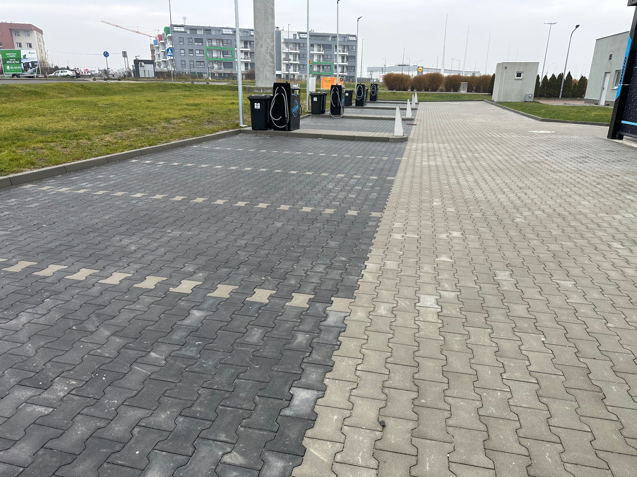 Ułożona kostka brukowa w dwóch odcieniach szarości, tworząca wzór na parkingu z odkurzaczami samochodowymi. Widoczne budynki w tle i elementy infrastruktury miejskiej.