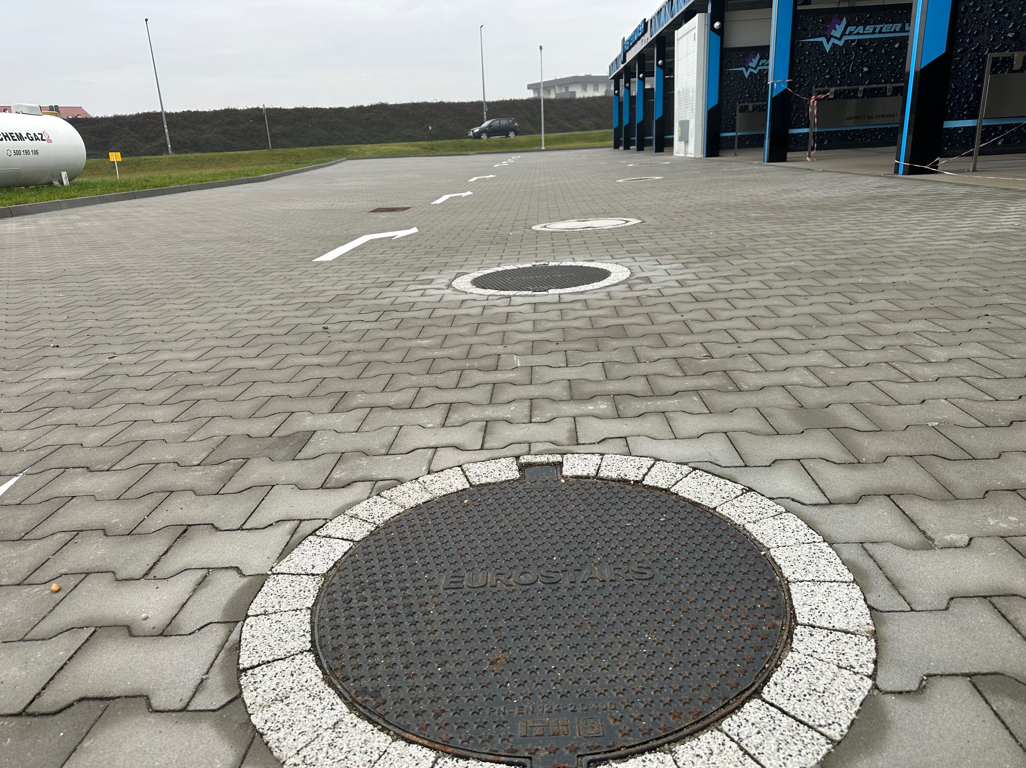 Szara kostka brukowa ułożona na parkingu z widocznymi studzienkami kanalizacyjnymi i białymi strzałkami kierunkowymi. W tle budynek myjni samochodowej.