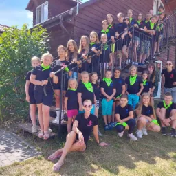 Grupa dzieci i dorosłych w czarnych koszulkach z logo i neonowo-zielonych chustach pozuje na schodach drewnianego budynku.