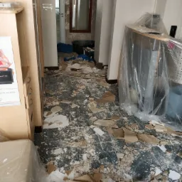 Wnętrze w trakcie remontu: gruz i resztki materiałów budowlanych na podłodze, meble zabezpieczone folią, widoczny fragment szafki i przejście do innego pomieszczenia.