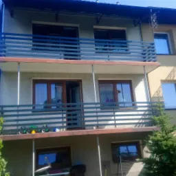 Dwupiętrowy dom z balkonami z poziomymi, szarymi balustradami, widok z dołu w słoneczny dzień.