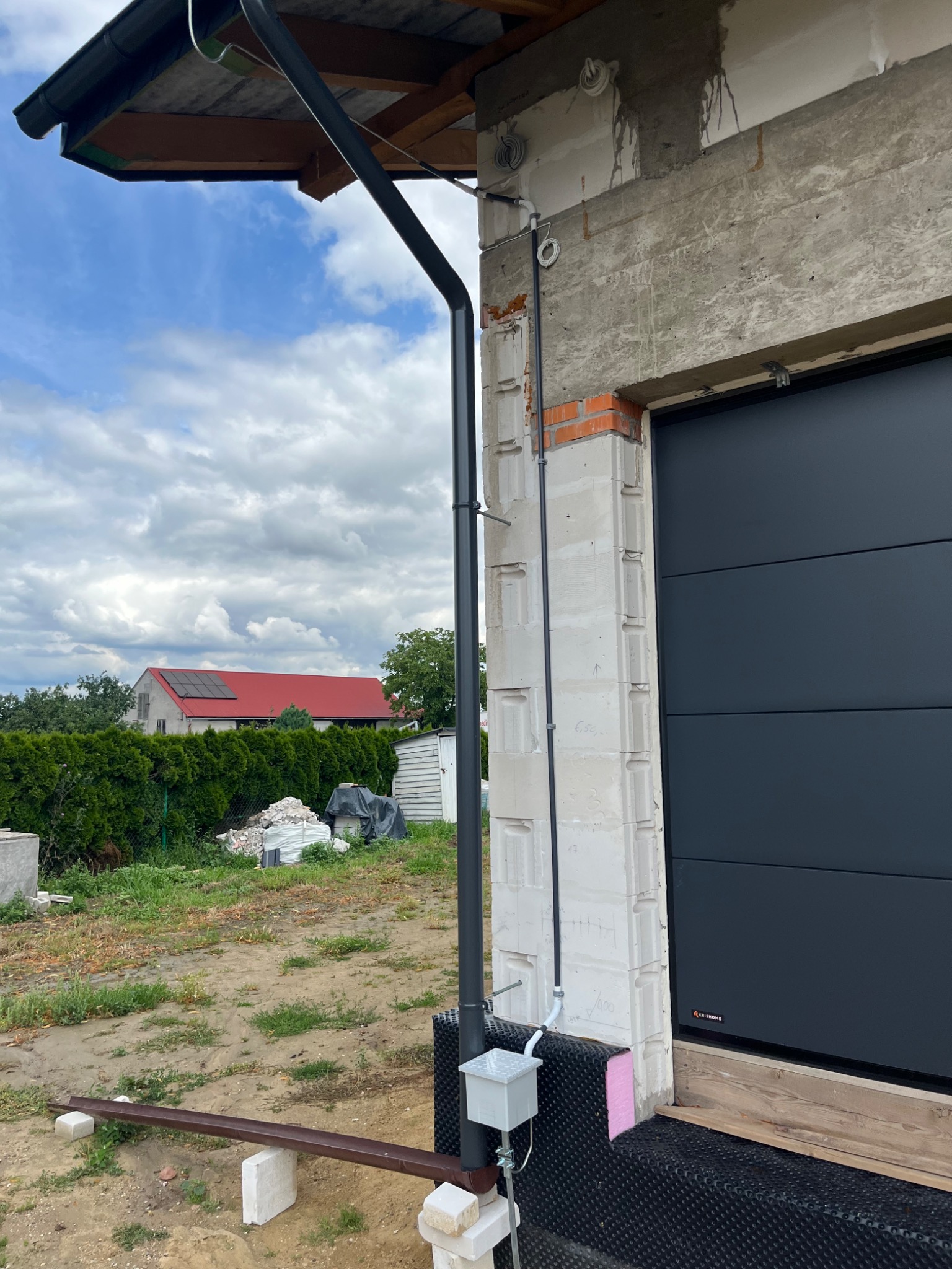 Zewnętrzna instalacja elektryczna przy garażu w trakcie budowy. Widoczne przewody, puszka elektryczna, fragment orynnowania i brama garażowa. W tle dom z czerwoną dachówką.
