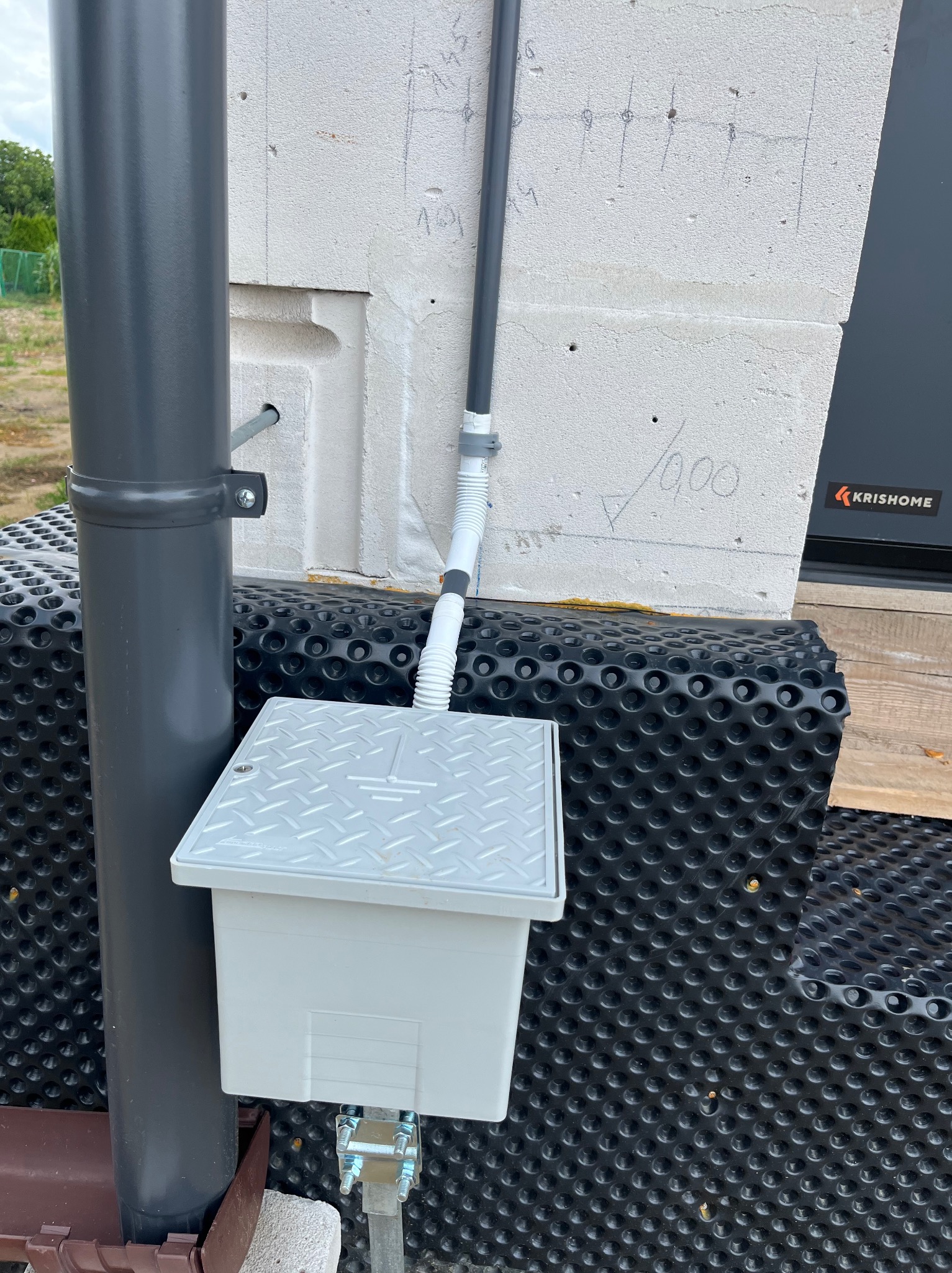 Szara skrzynka elektryczna z metalową pokrywą, przymocowana do ściany budynku z izolacją, z widocznym przewodem i rurą spustową. Ślady ołówka na ścianie.