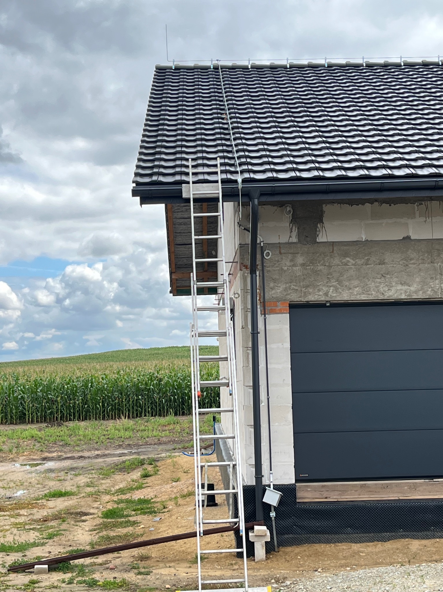 Montaż instalacji odgromowej na dachu budynku z szarą dachówką, widoczna drabina oparta o ścianę. W tle pole kukurydzy i pochmurne niebo.