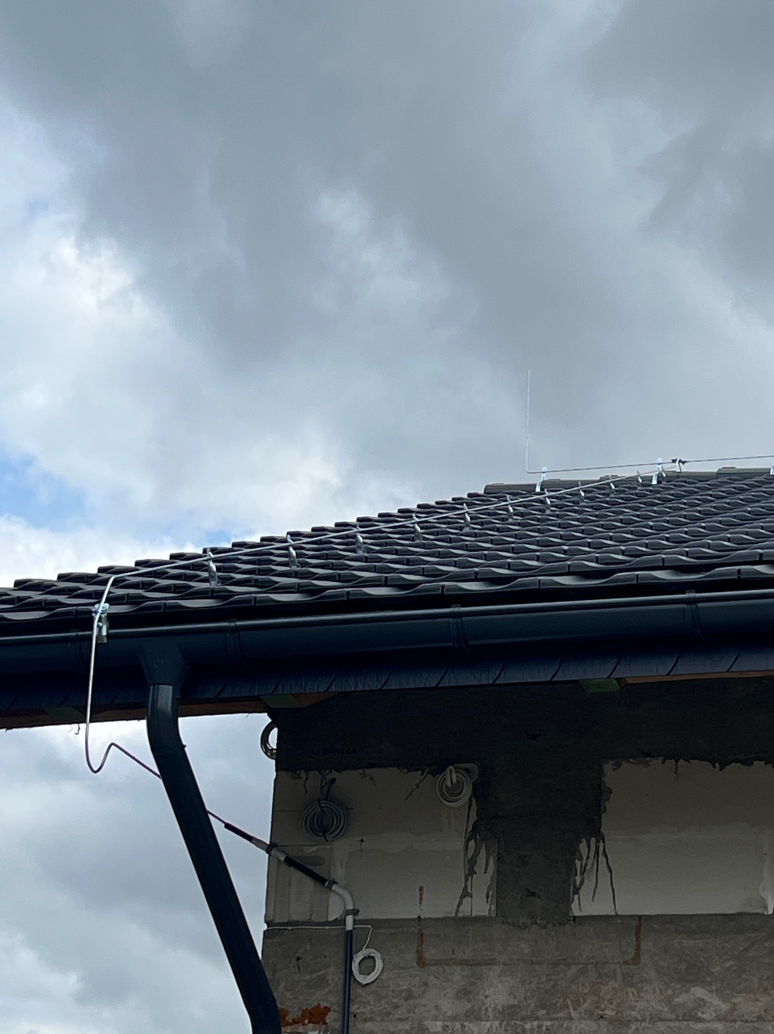 Instalacja odgromowa na dachu z czarnej dachówki, widoczne przewody i uchwyty mocujące. Budynek w stanie surowym, widoczne przewody elektryczne.
