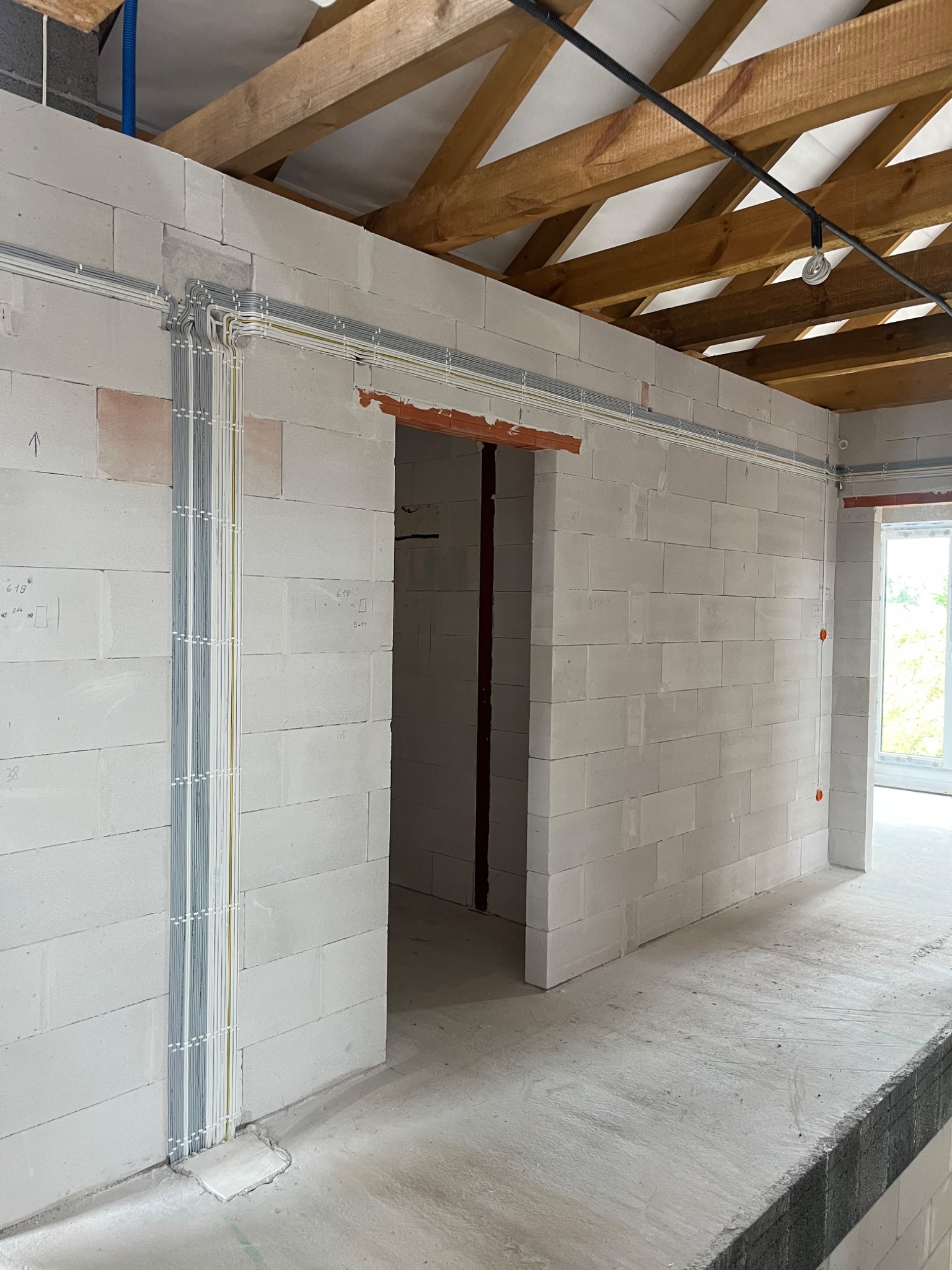 Wnętrze w trakcie instalacji elektrycznej: kable w listwach na ścianach z betonu komórkowego, drewniany strop. Widoczne okablowanie i punkty elektryczne.