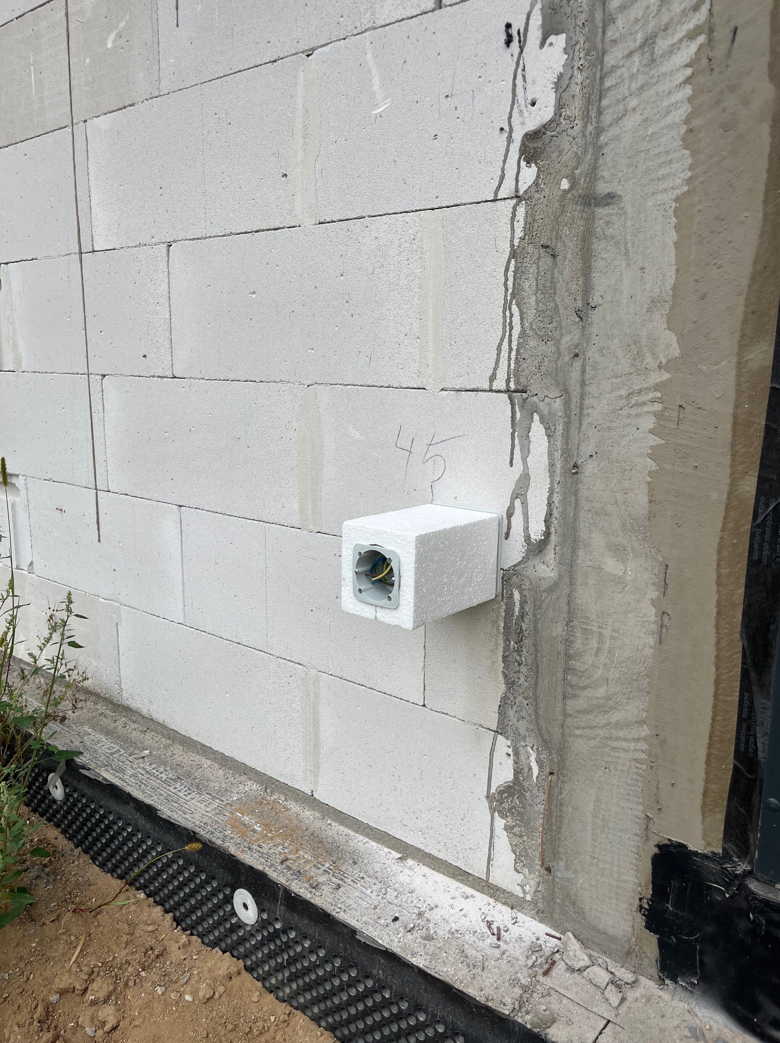 Biała puszka elektryczna wystająca ze ściany z betonu komórkowego. Widoczne przewody. Na ścianie numer '45' napisany ołówkiem. Izolacja fundamentów przy ziemi.