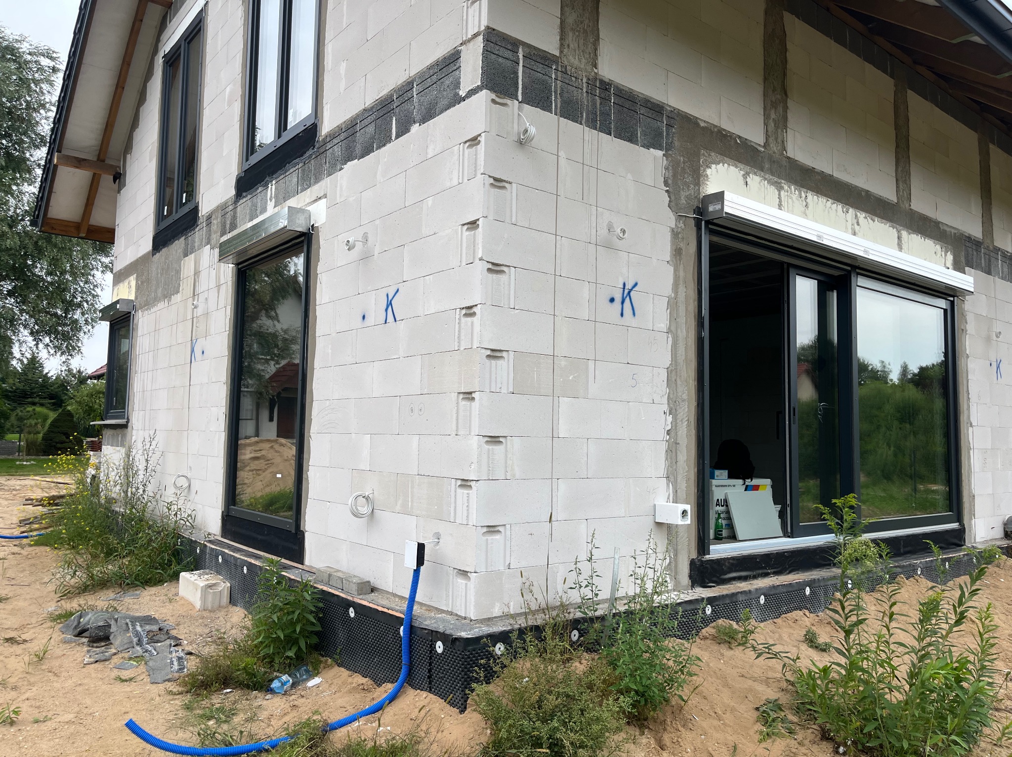 Surowy stan budynku z białego pustaka z zamontowanymi oknami i roletami zewnętrznymi. Widoczne wystające przewody elektryczne i niebieski peszel.