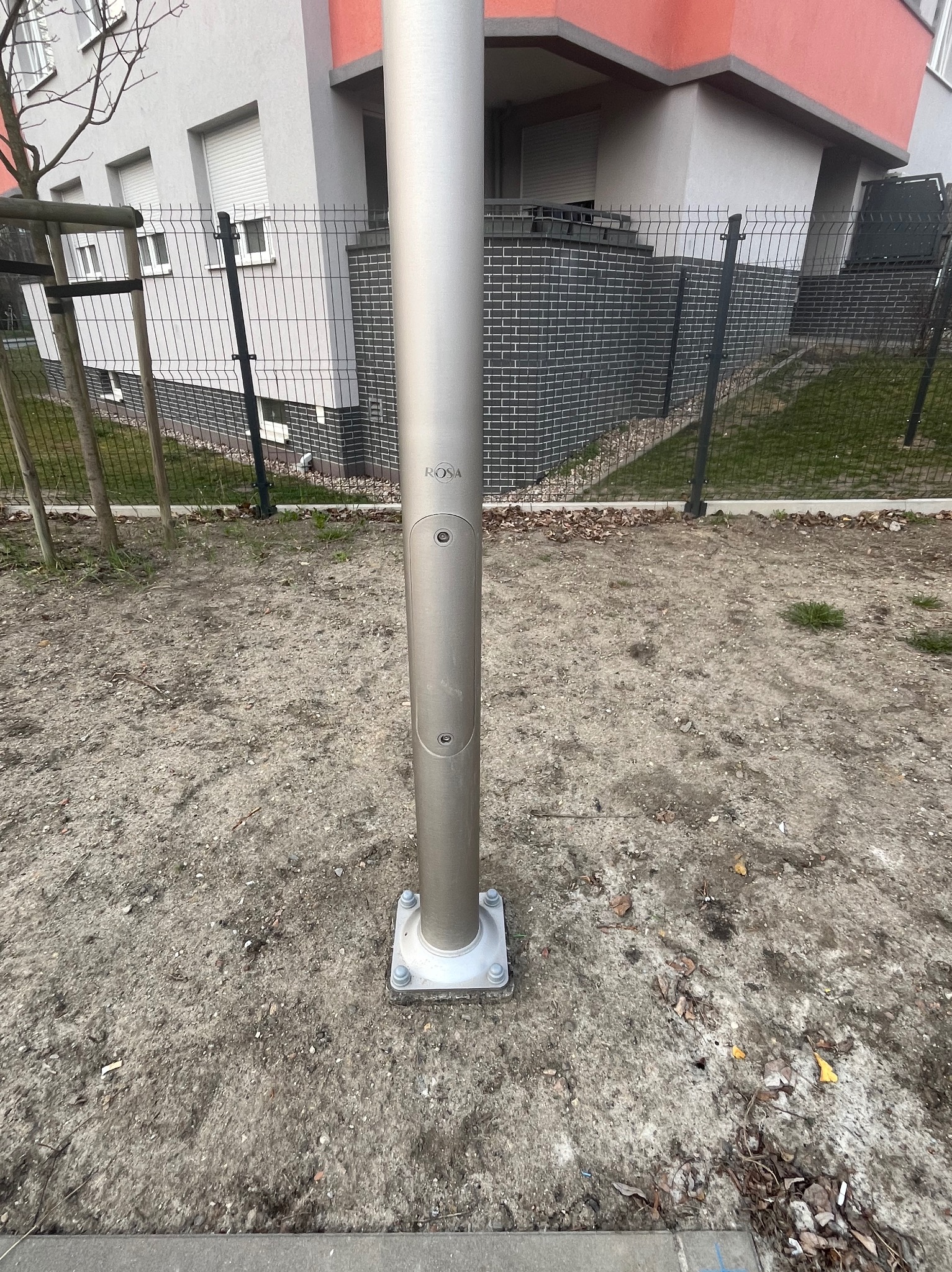 Metalowa instalacja odgromowa marki Rosa, zamontowana na fundamencie śrubami, na tle ogrodzenia i budynku mieszkalnego.