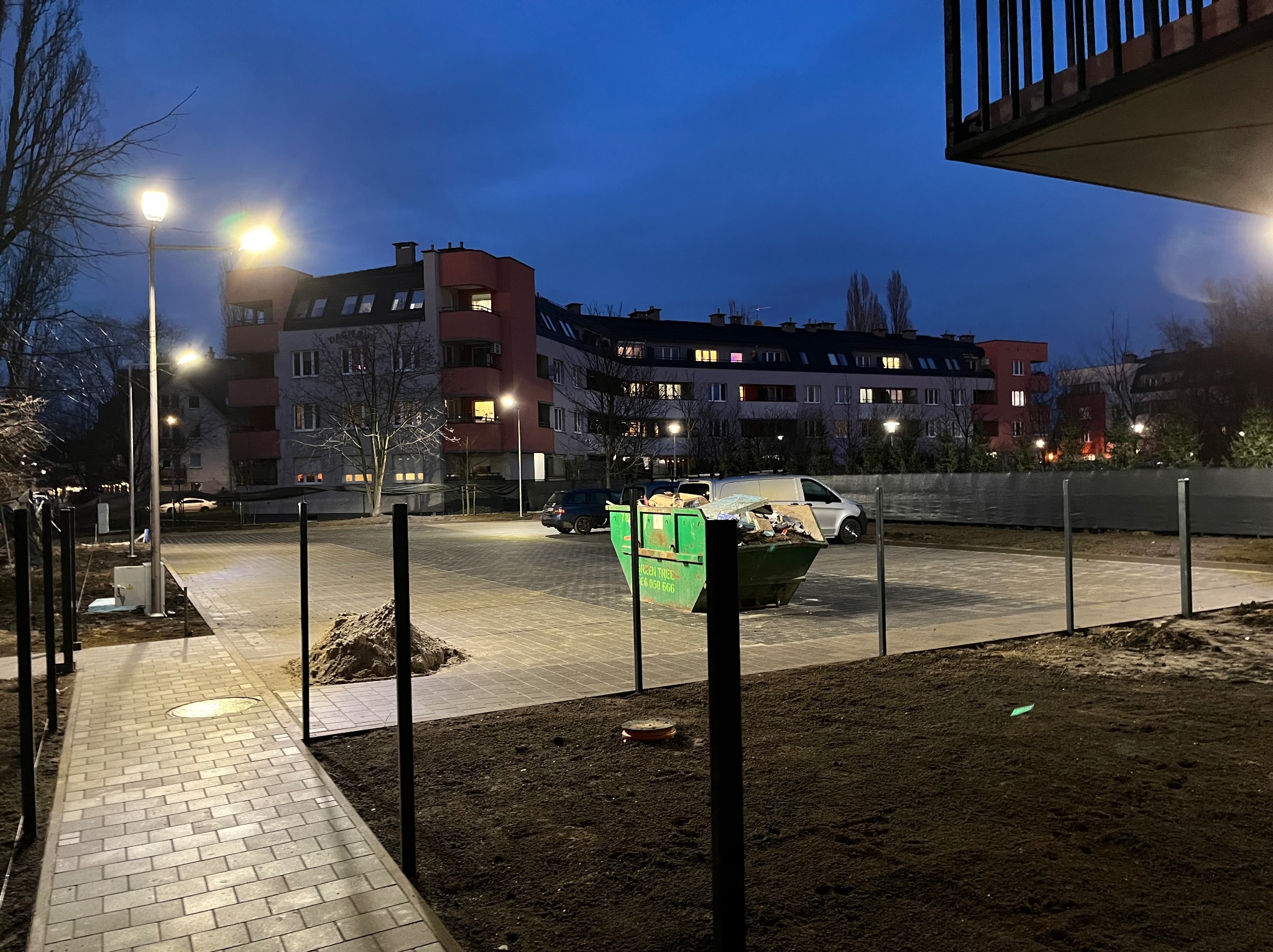 Wieczorny widok na plac budowy z kontenerem na gruz, nową kostką brukową i słupami oświetleniowymi na tle bloku mieszkalnego.
