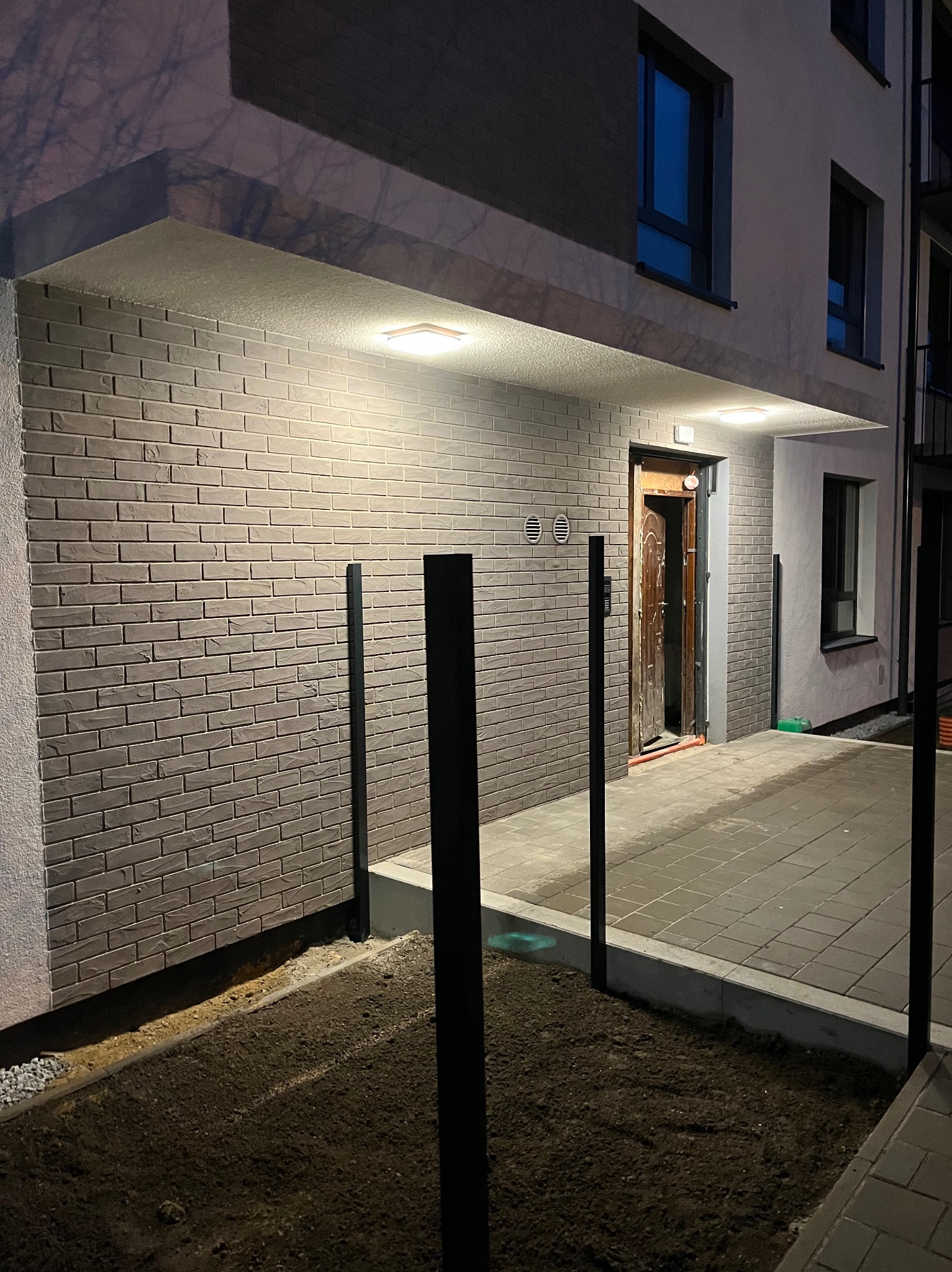 Oświetlony wjazd do budynku z ceglaną elewacją, widoczne czarne słupki ogrodzenia i fragment niedokończonego trawnika. Nocne ujęcie.