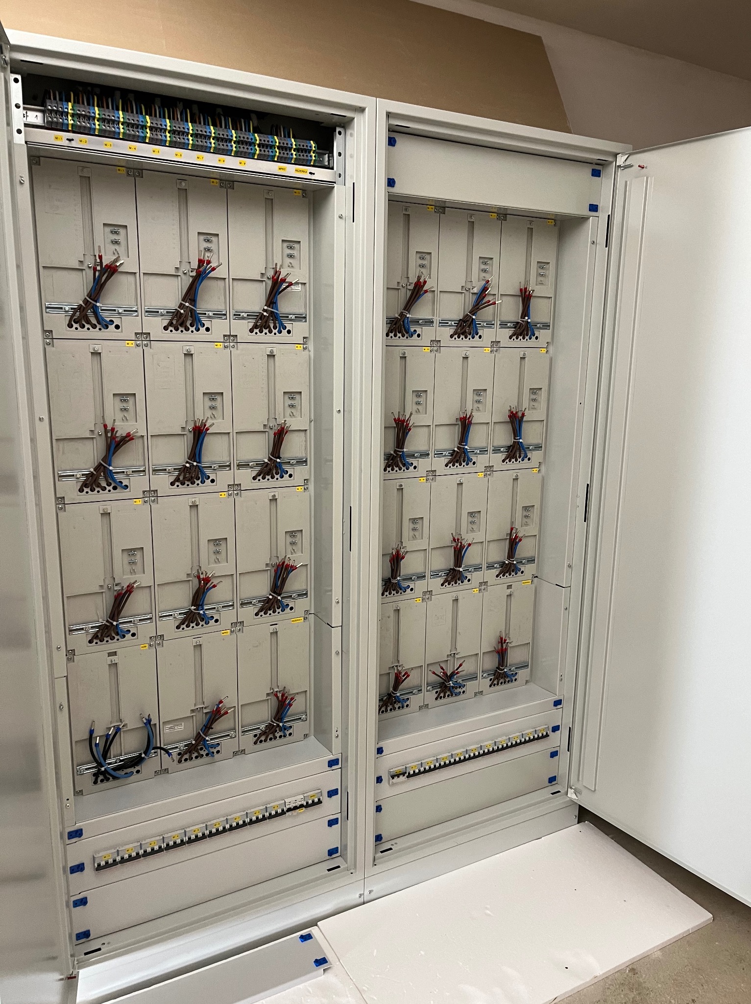 Otwarta, duża szafa elektryczna z licznymi modułami i przewodami. Widoczne złącza, bezpieczniki i kable w kolorach: czerwonym, niebieskim i czarnym. Szara obudowa.