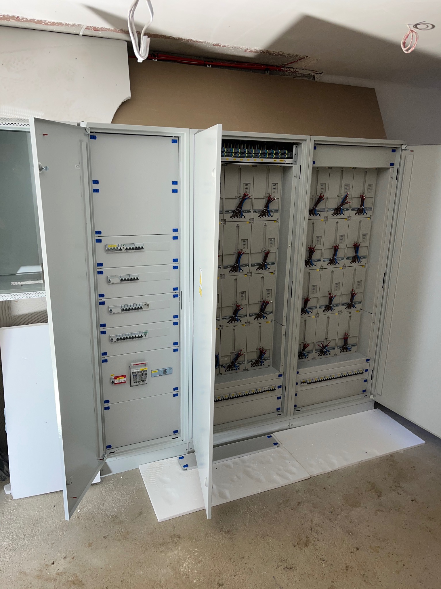 Rozdzielnica elektryczna z otwartymi drzwiami, widoczne zabezpieczenia i okablowanie. Biała obudowa, surowy stan wykończenia wnętrza, widoczne kable pod sufitem.