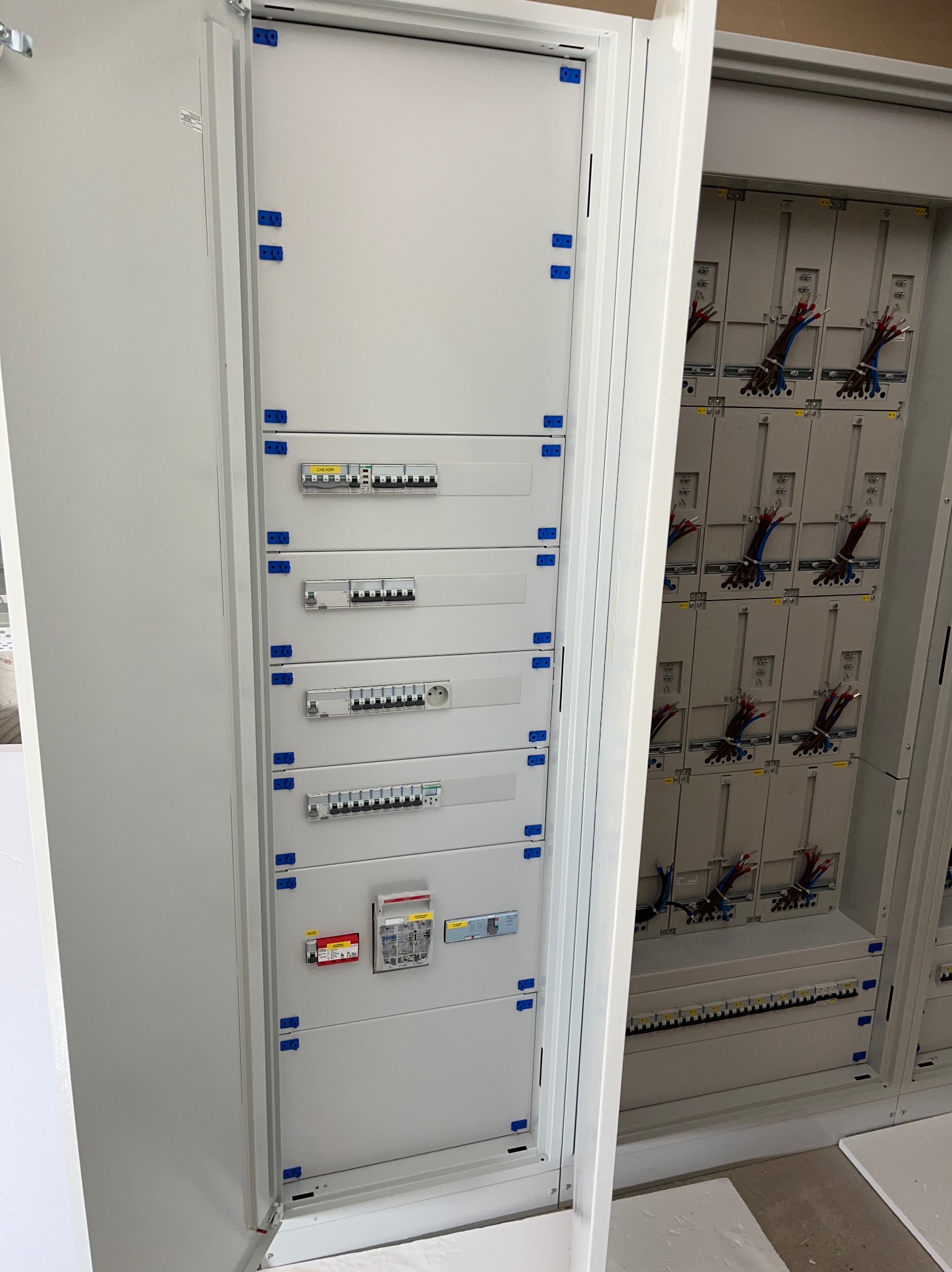 Rozdzielnia elektryczna z bezpiecznikami i przewodami w szafie, widok z bliska na zamontowane elementy. Biała obudowa, niebieskie mocowania.