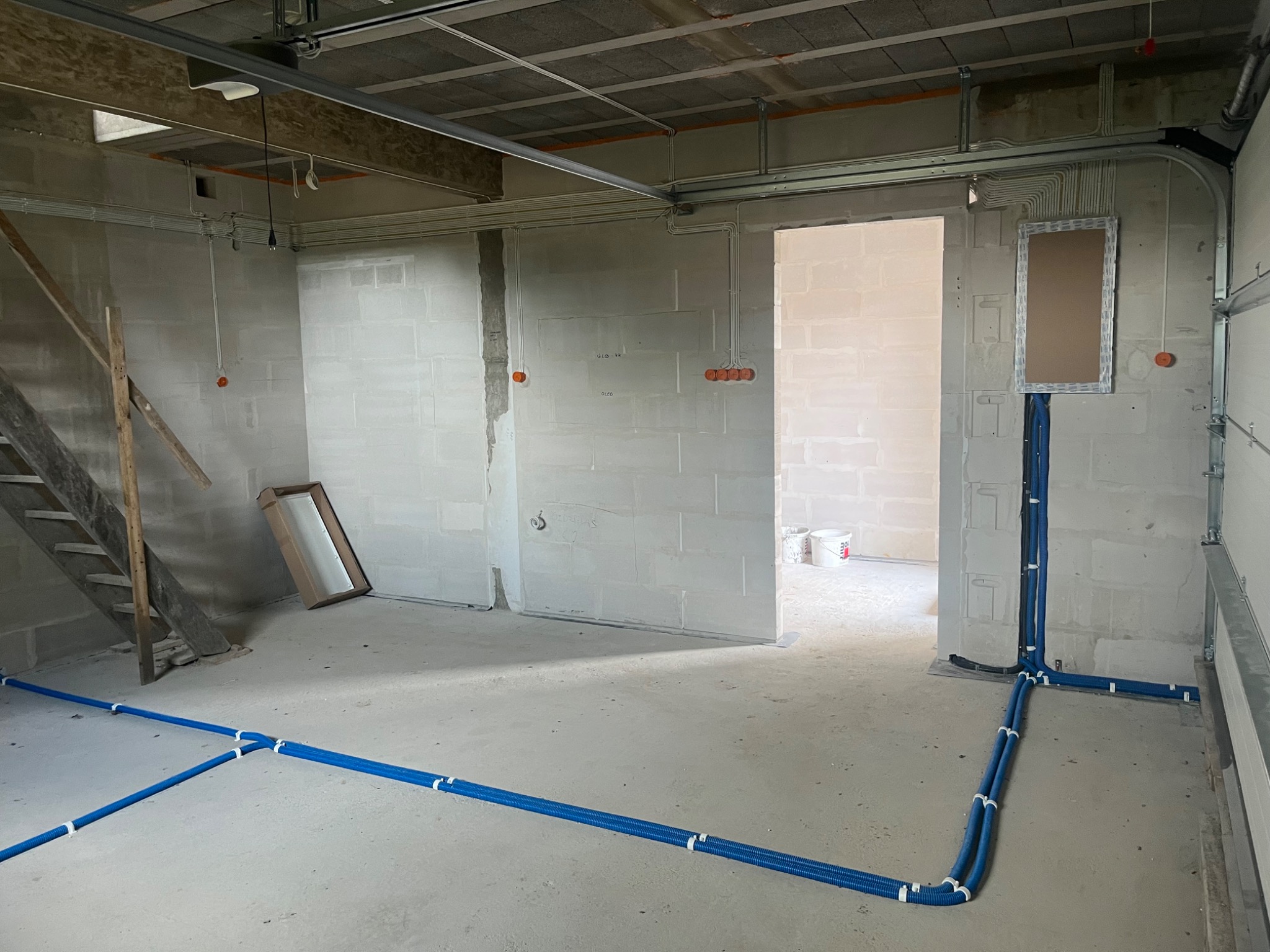 Surowy garaż z widoczną, częściowo rozprowadzoną instalacją elektryczną w peszlach na ścianach i podłodze. Widoczne puszki elektryczne i schody.