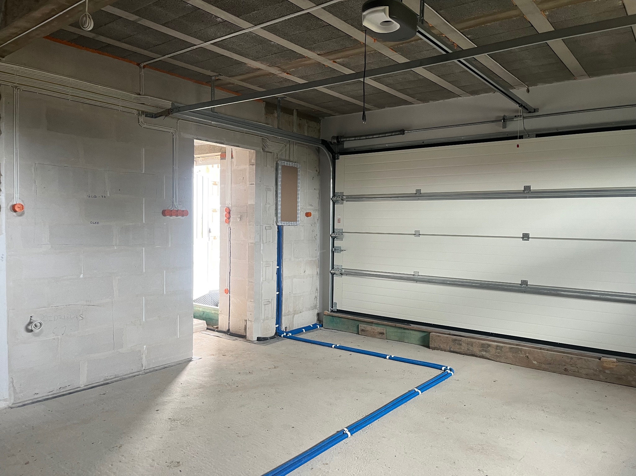 Surowy garaż z widoczną instalacją elektryczną w peszlu, z puszkami na ścianach i skrzynką rozdzielczą. Widoczna brama garażowa i sufit z betonu komórkowego.