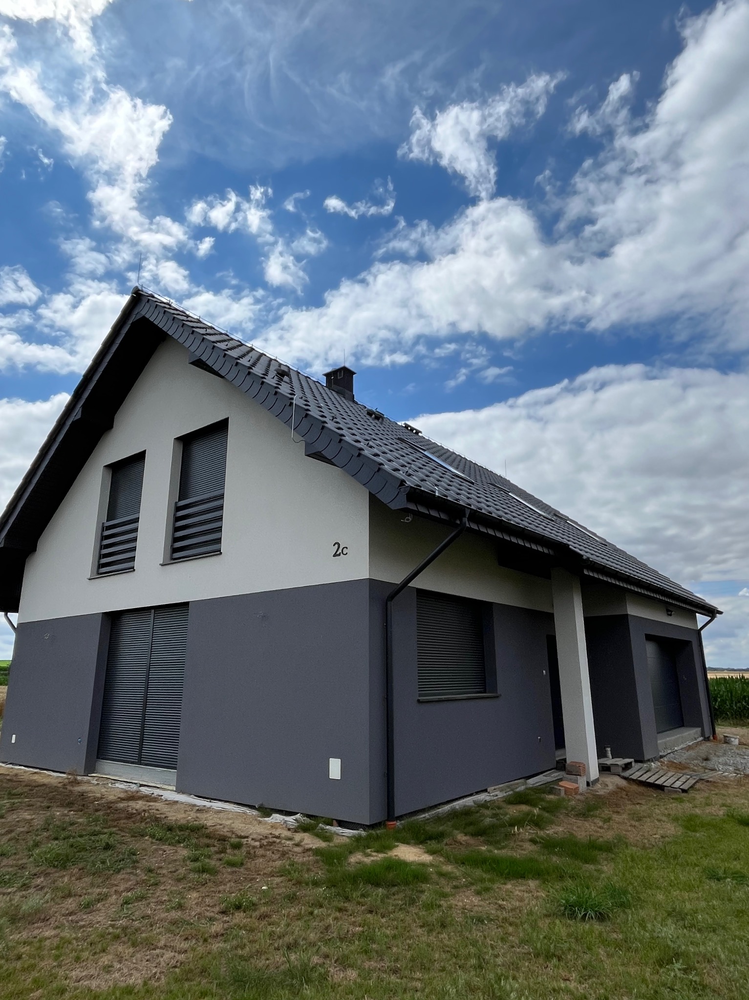 Dwukondygnacyjny dom z szarą elewacją i ciemną dachówką na tle błękitnego nieba z chmurami. Widoczne rolety zewnętrzne w oknach.