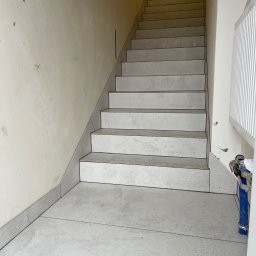 DB Dawid Bojarski - Wyłożone szarymi płytkami schody z ciemnymi fugami, ściana w trakcie przygotowania pod malowanie, widoczny grzejnik i akcesoria instalacyjne