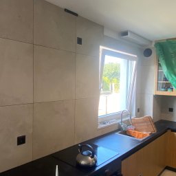 DB Dawid Bojarski - Kuchnia w trakcie wykańczania: ściany wyłożone płytami imitującymi beton, czarny blat, stalowy czajnik na płycie indukcyjnej, okno z widokiem na zieleń, szafki częściowo zakryte folią ochronną.