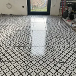 Ułożona podłoga z błyszczących, czarno-białych płytek w geometryczny wzór, widoczne niedokończone ściany i narzędzia glazurnicze w tle.