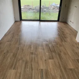 Świeżo ułożona, połyskująca podłoga z płytek imitujących drewno w pustym pomieszczeniu z dużym oknem balkonowym i widokiem na podwórze z ułożonymi deskami; dolna krawędź wykończona dekoracyjną...