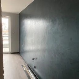 Ściana w pokoju pokryta dekoracyjną, ciemną farbą z efektem struktury, widoczne puszki instalacyjne i listwy przypodłogowe przed montażem, parkiet ułożony.