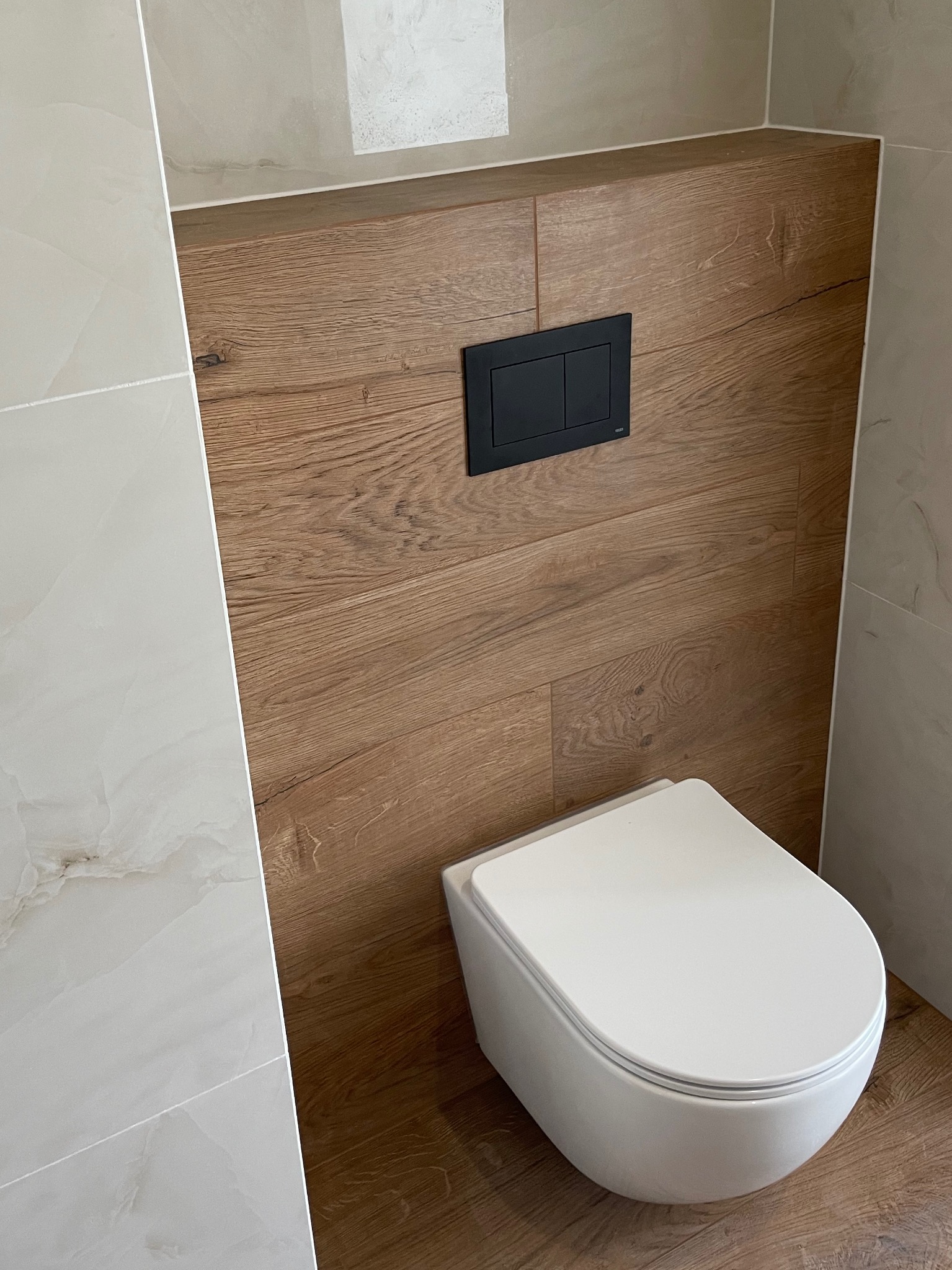 Nowoczesna toaleta podwieszana z czarnym panelem sterującym na ściance obłożonej panelami drewnopodobnymi, obok ściana wyłożona białymi płytkami imitującymi marmur.