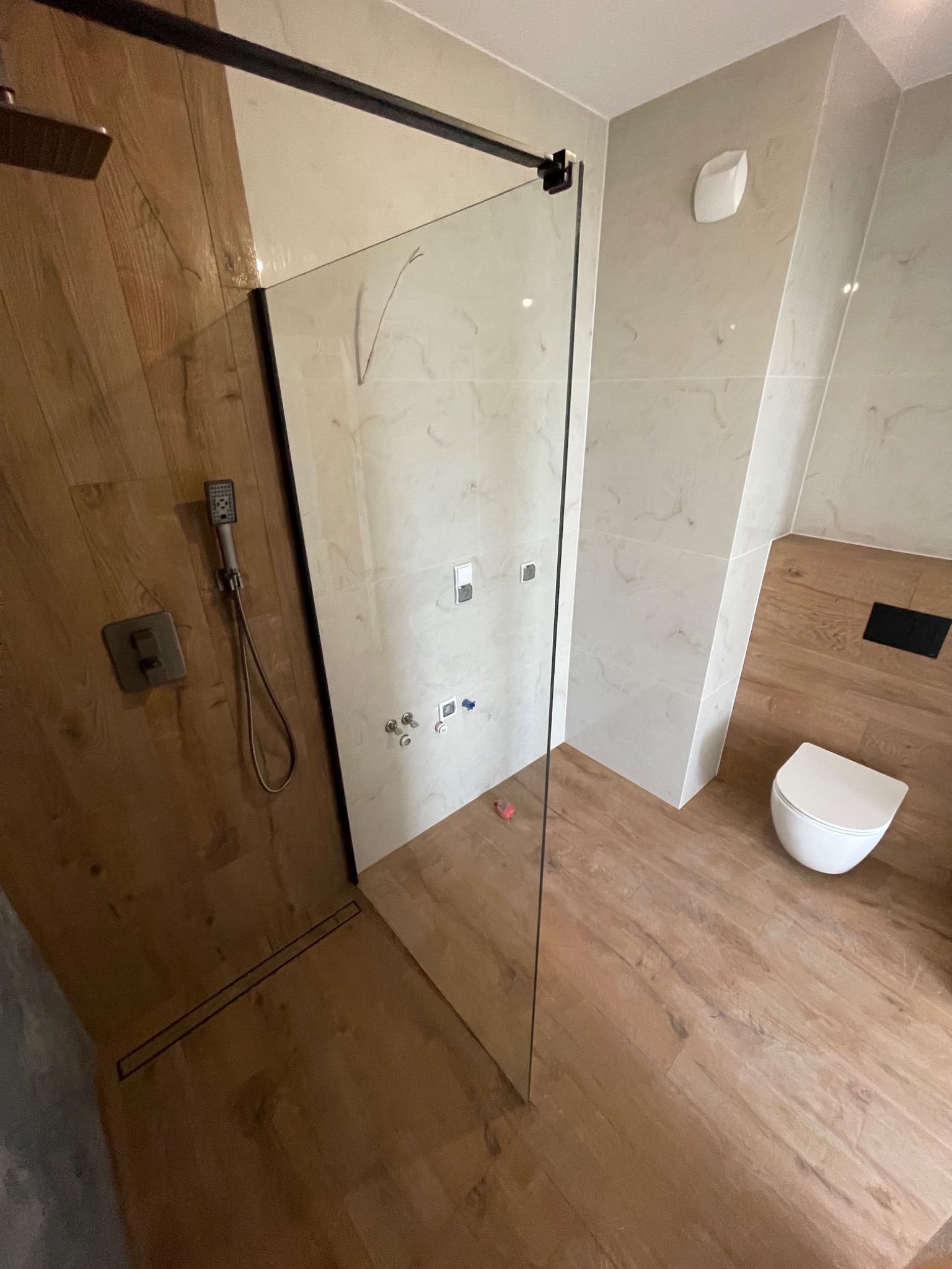 Nowoczesna kabina prysznicowa typu walk-in z czarnymi profilami, drewnopodobne płytki na ścianie prysznica kontrastujące z marmurowymi płytkami na pozostałych ścianach, wisząca miska WC, odpływ...