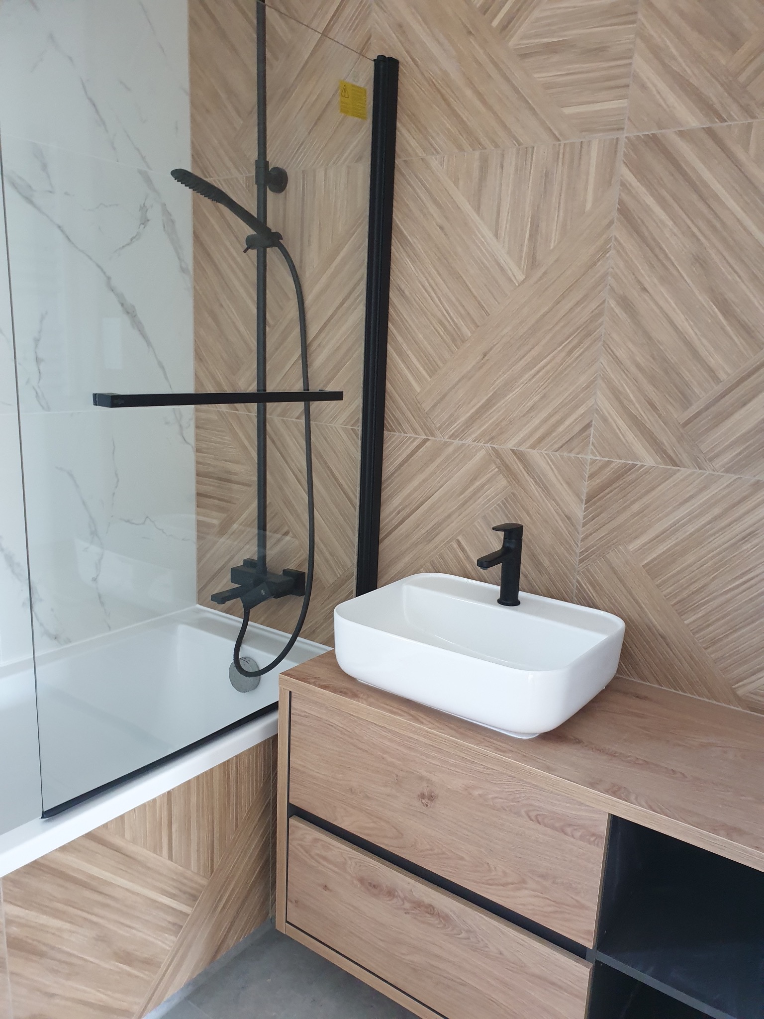 Nowoczesna łazienka w Gdyni po remoncie. Na pierwszym planie biała umywalka nablatowa z czarną armaturą, obok kabina prysznicowa z czarnymi profilami i deszczownicą. Na ścianie i obudowie wanny...