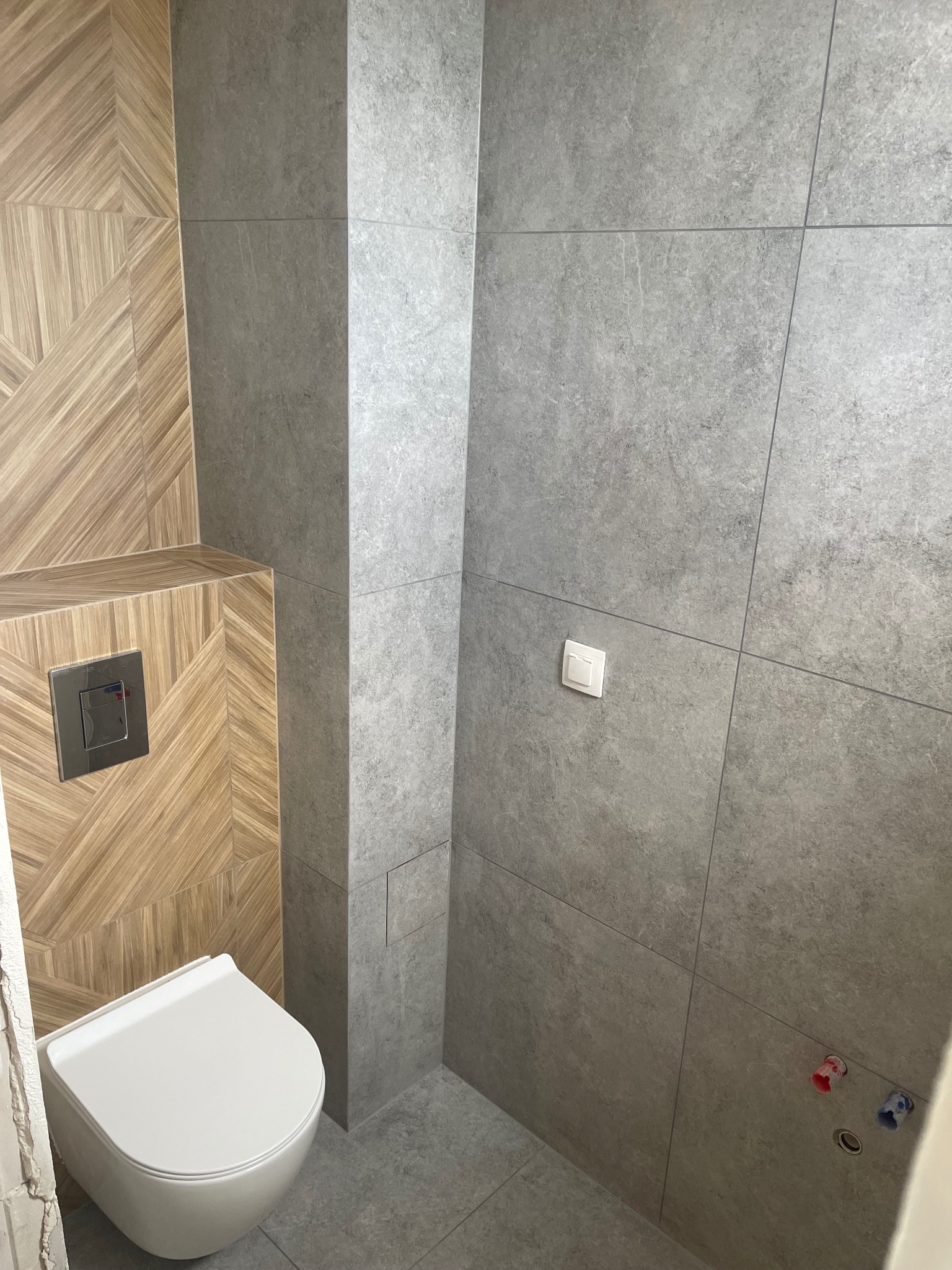 Wykończona łazienka z drewnopodobnymi płytkami ułożonymi w geometryczny wzór na ścianie za podwieszaną toaletą i szarymi płytkami imitującymi kamień na pozostałych ścianach, widoczny chromowany...