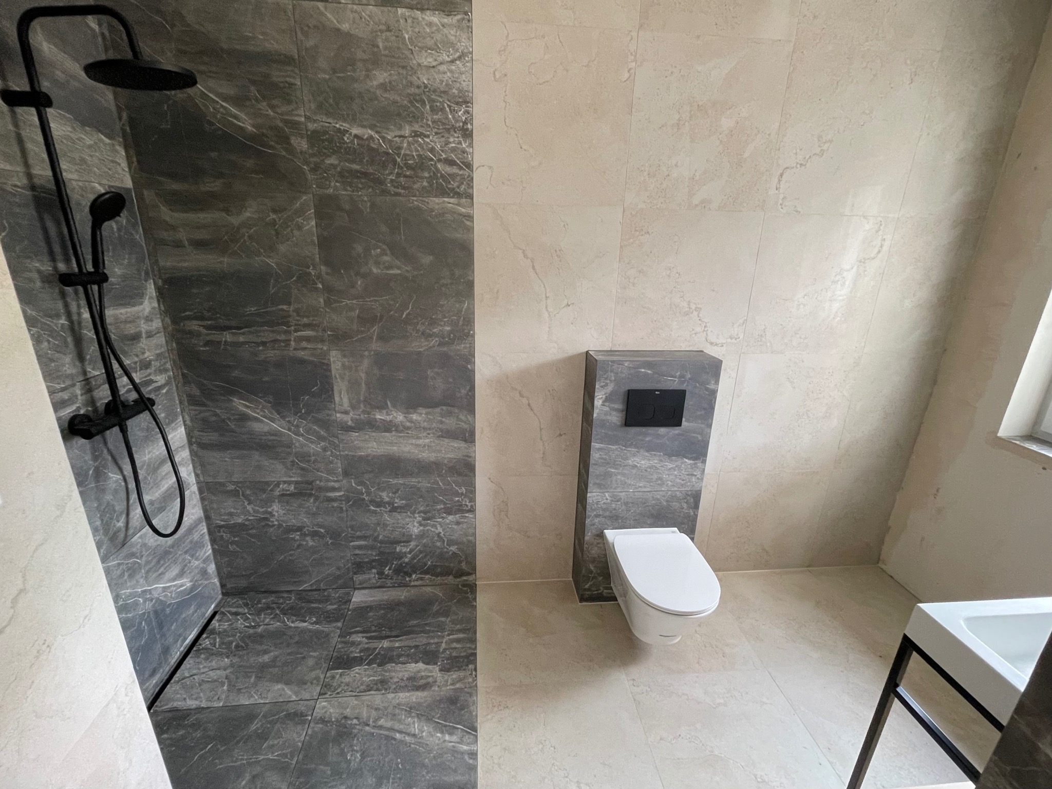 Nowoczesna łazienka z kabiną prysznicową typu walk-in wyłożoną szarymi płytkami imitującymi marmur, kontrastującymi z jasnymi płytkami na pozostałej powierzchni. Czarna armatura prysznicowa...