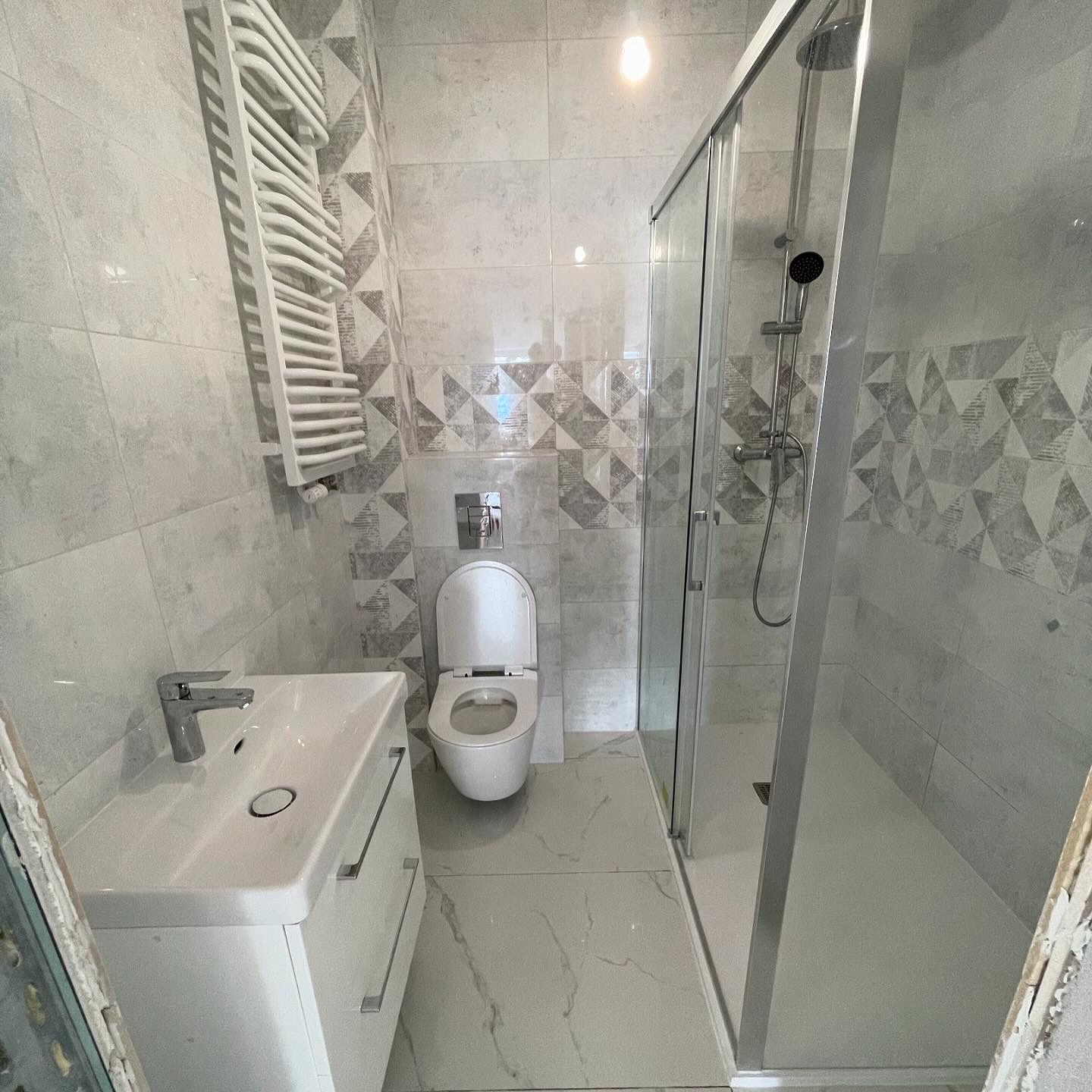 Jasna łazienka z kabiną prysznicową, umywalką z szafką, toaletą i grzejnikiem drabinkowym, wykończona płytkami w odcieniach szarości z geometrycznym wzorem na ścianie za toaletą.