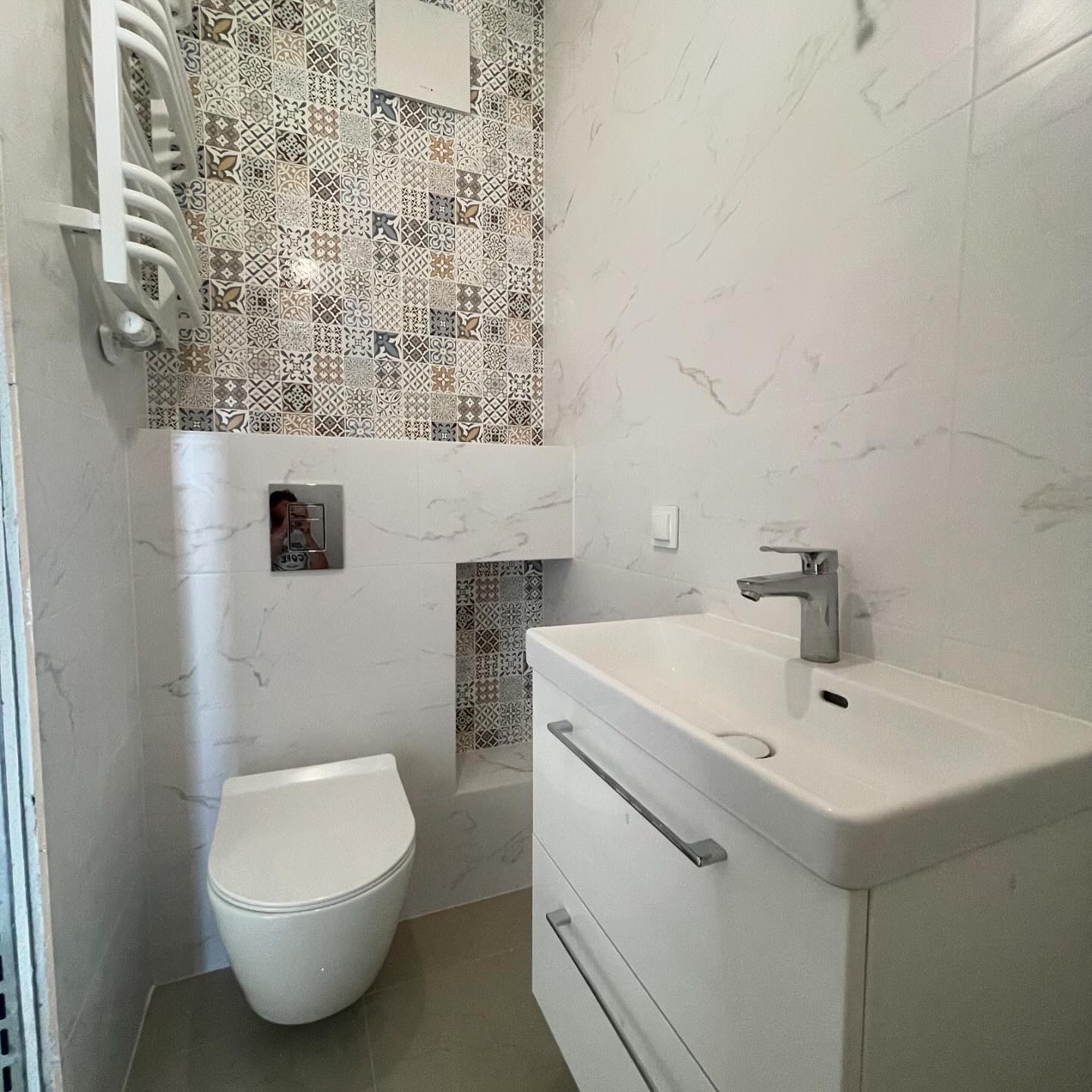 Nowoczesna łazienka z patchworkową ścianą z płytek, białą umywalką z szafką i podwieszaną toaletą.