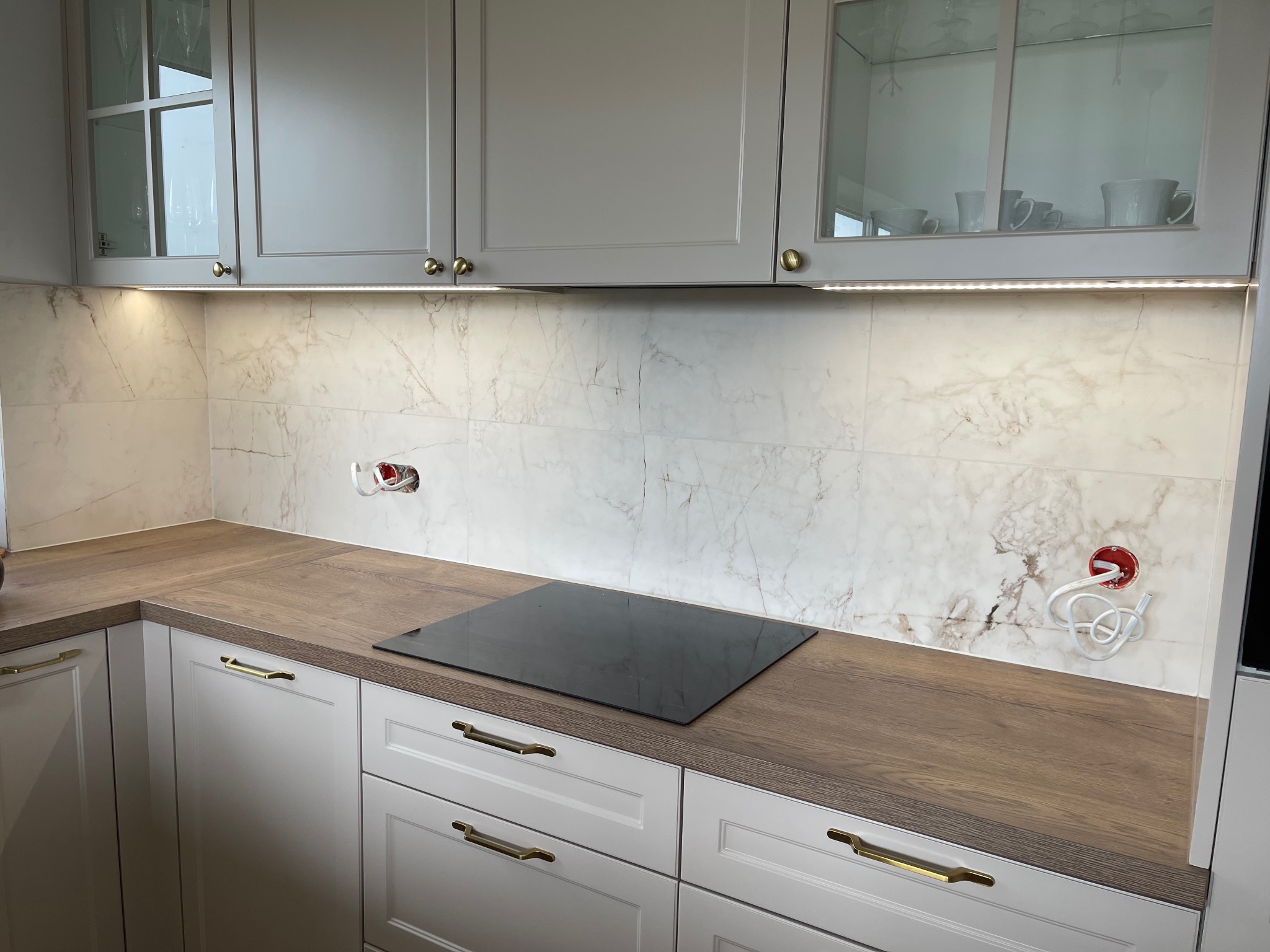 Wykończona kuchnia z marmurową ścianą, drewnianym blatem, indukcyjną płytą i szarymi szafkami z oświetleniem LED podszafkowym, widoczne gniazdka elektryczne przed podłączeniem.