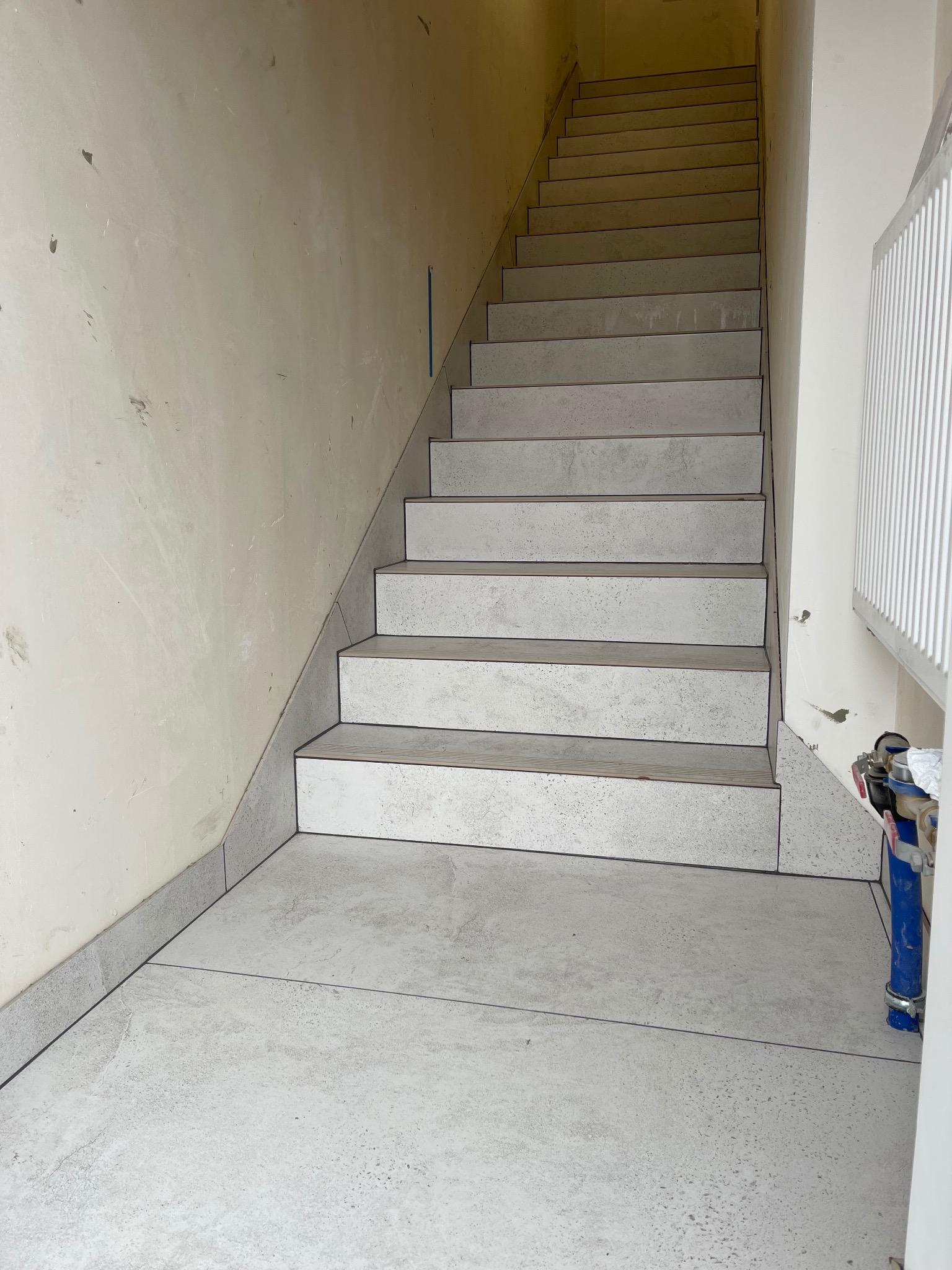 Wyłożone szarymi płytkami schody z ciemnymi fugami, ściana w trakcie przygotowania pod malowanie, widoczny grzejnik i akcesoria instalacyjne