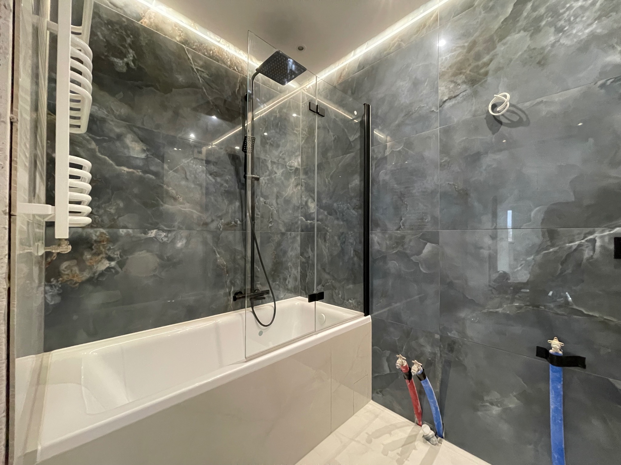 Nowoczesna łazienka z wanną obudowaną białymi płytkami, szklanym parawanem, deszczownicą i grzejnikiem drabinkowym na tle płytek imitujących marmur w odcieniach szarości, z podświetleniem LED.