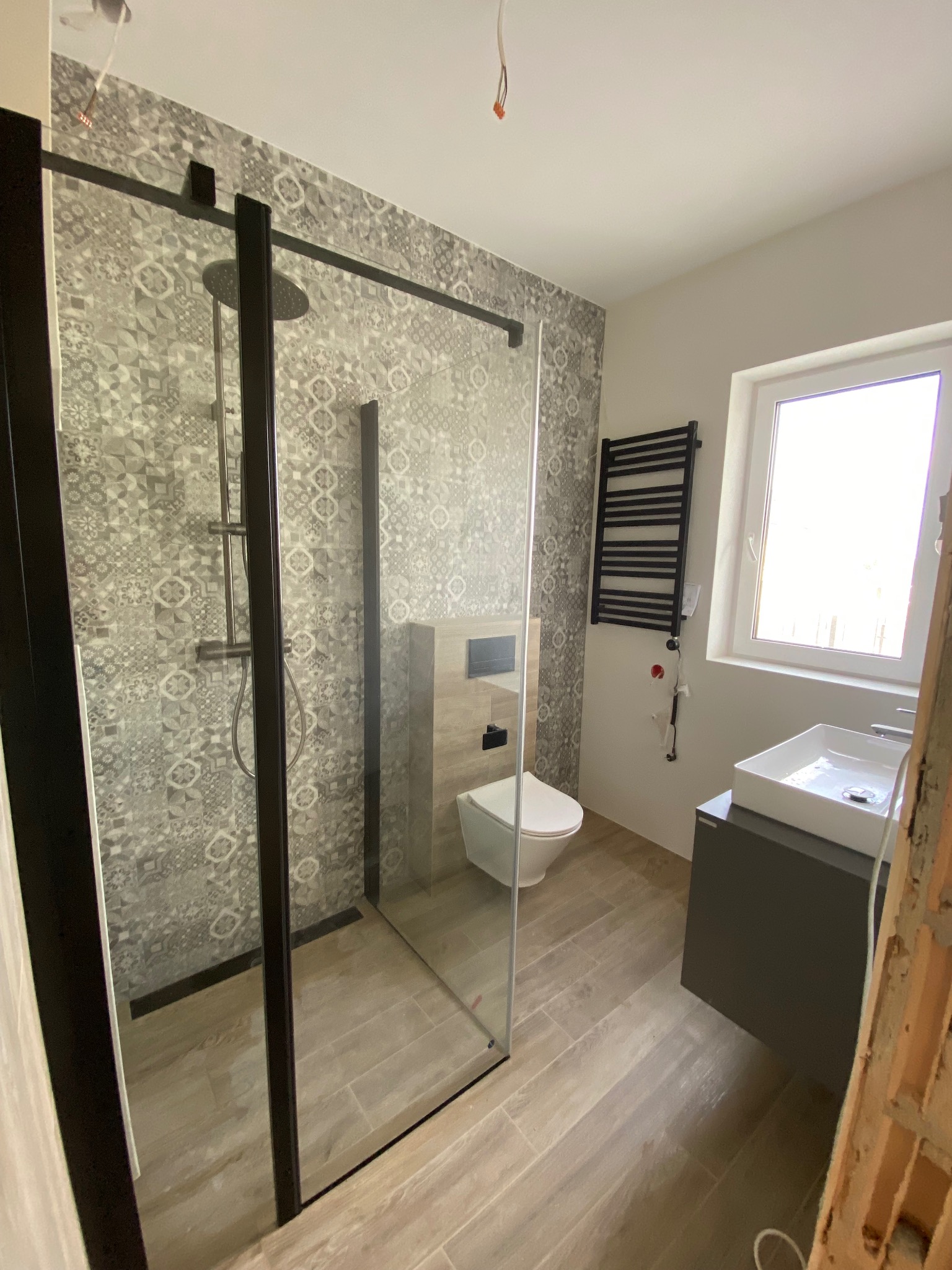 Nowoczesna łazienka z kabiną prysznicową typu walk-in z czarnymi profilami, wzorzystymi płytkami na ścianie prysznica, podwieszaną toaletą i grzejnikiem drabinkowym.