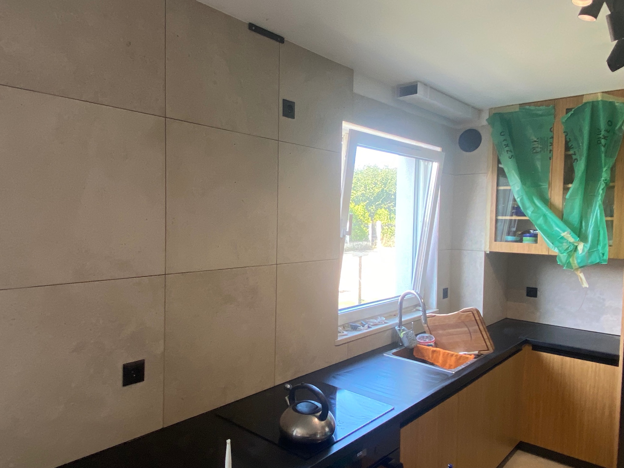 Kuchnia w trakcie wykańczania: ściany wyłożone płytami imitującymi beton, czarny blat, stalowy czajnik na płycie indukcyjnej, okno z widokiem na zieleń, szafki częściowo zakryte folią ochronną.