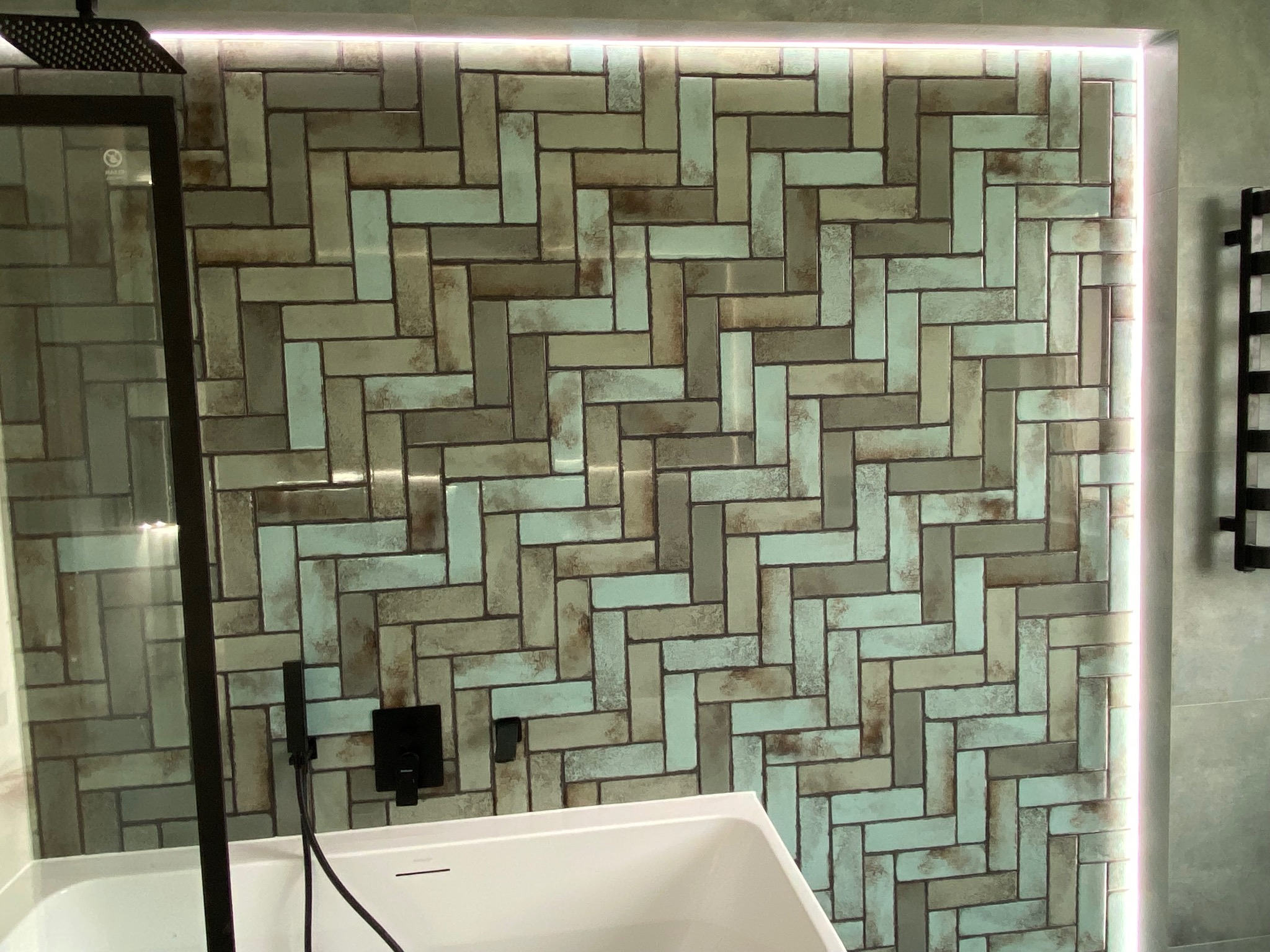 Ściana w łazience wyłożona prostokątnymi, błyszczącymi płytkami w odcieniach szarości, brązu i błękitu ułożonymi w jodełkę, z czarną armaturą i fragmentem białej wanny, oświetlona listwą LED.