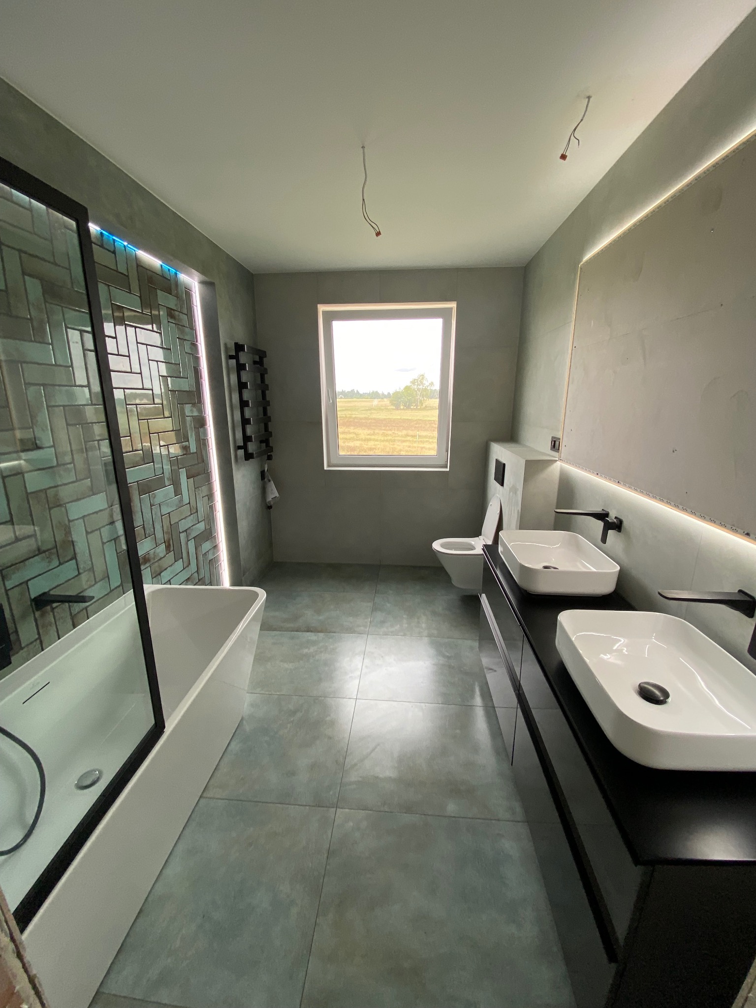 Nowoczesna łazienka w szaro-zielonej tonacji z wanną za szklaną ścianką, podwójną umywalką na czarnym blacie, grzejnikiem drabinkowym i oknem z widokiem na pole.