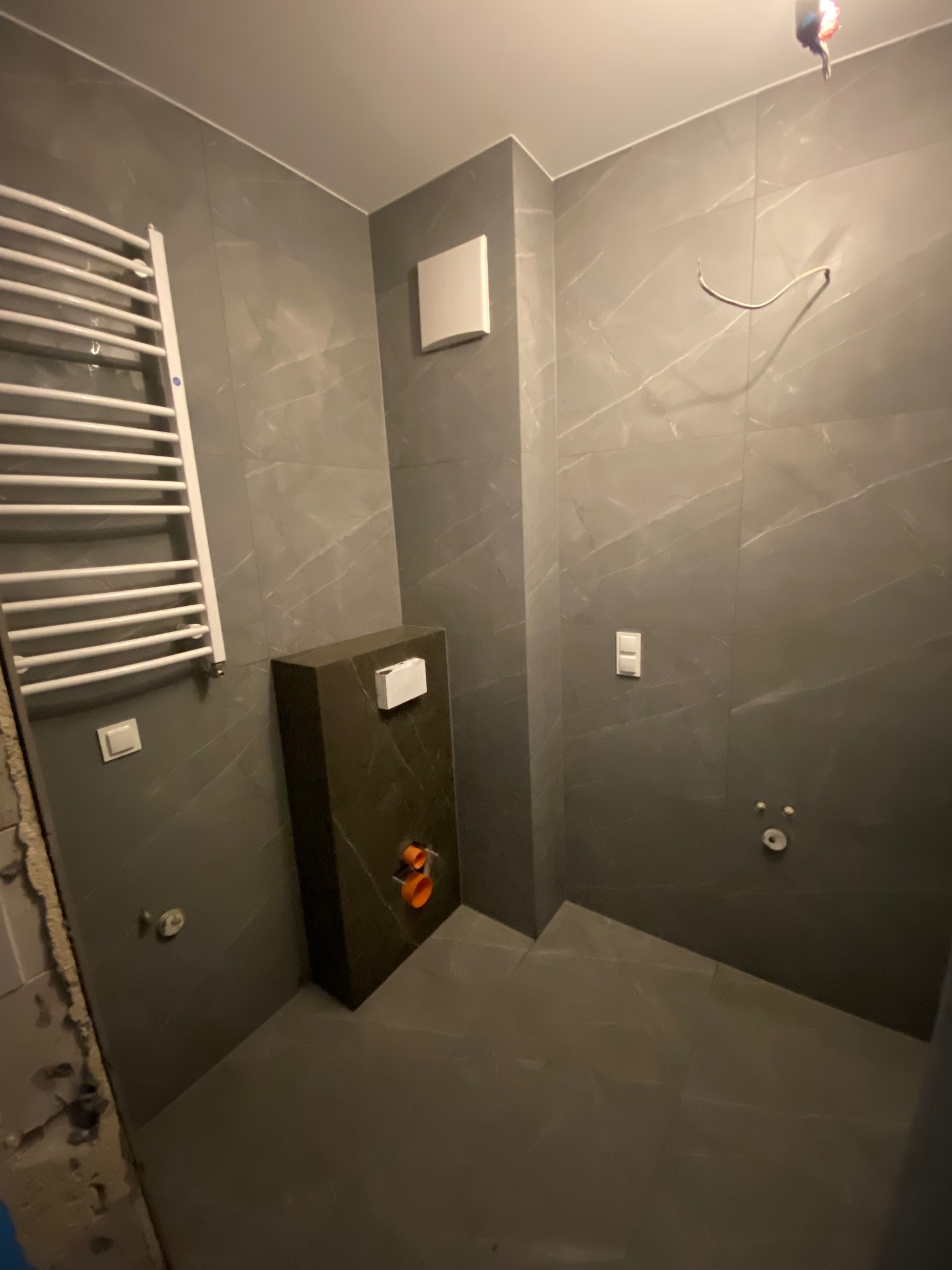 Szara łazienka w trakcie wykończenia, widoczne płytki imitujące marmur, grzejnik drabinkowy, zabudowa WC z przyciskiem, instalacja elektryczna i hydrauliczna przed podłączeniem armatury.